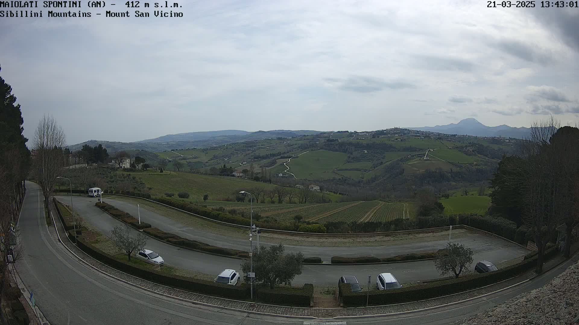 A mostly cloudy day reveals a winding road leading to a parking lot overlooking a hilly landscape of vineyards and distant mountains.