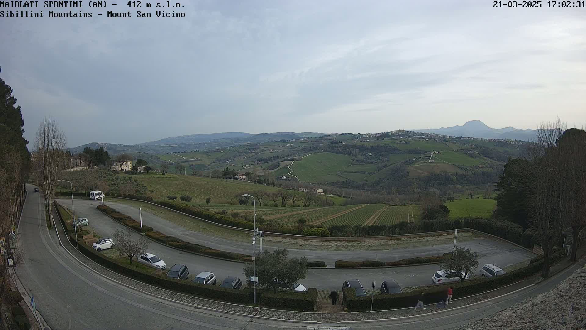 A mostly cloudy day reveals a hilly landscape with vineyards and a parking lot containing several cars, overlooked by a road curving through the scene.