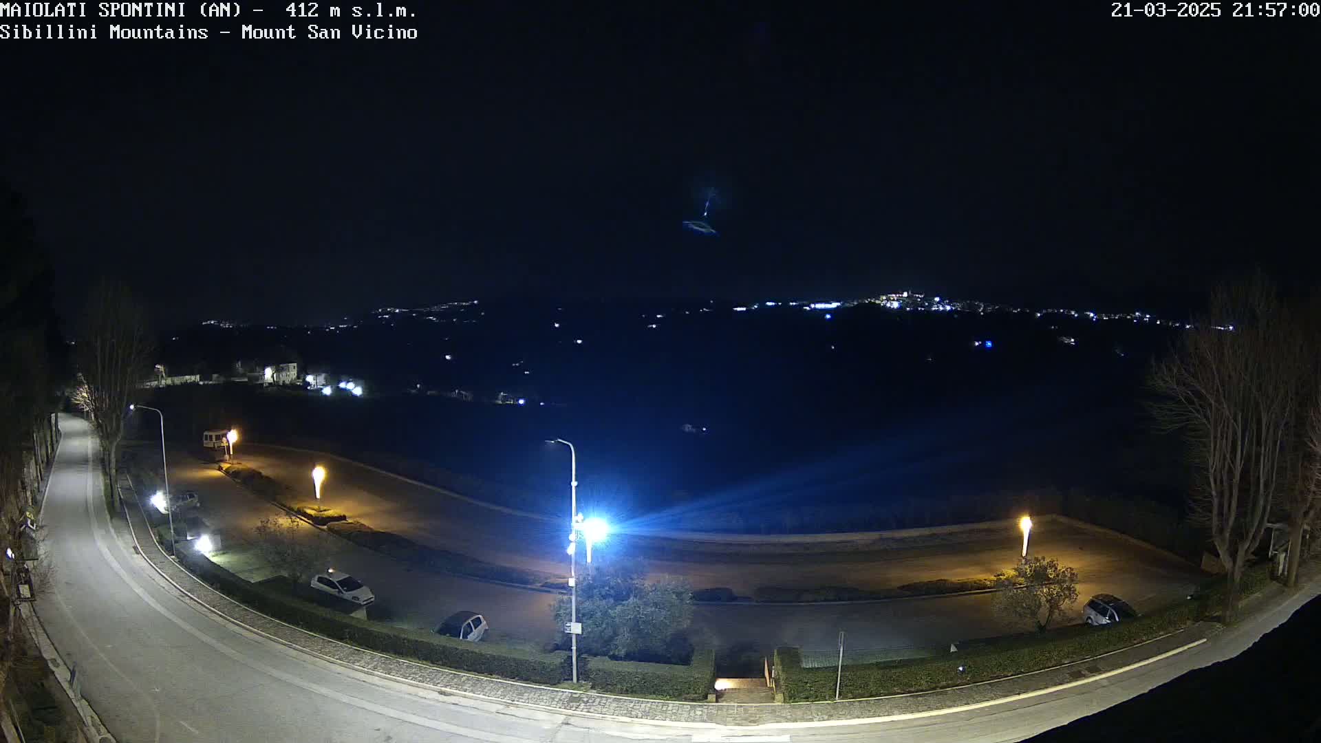 A nighttime view of a curving road with parked cars and streetlights, overlooking a distant, dimly lit town under a clear sky.