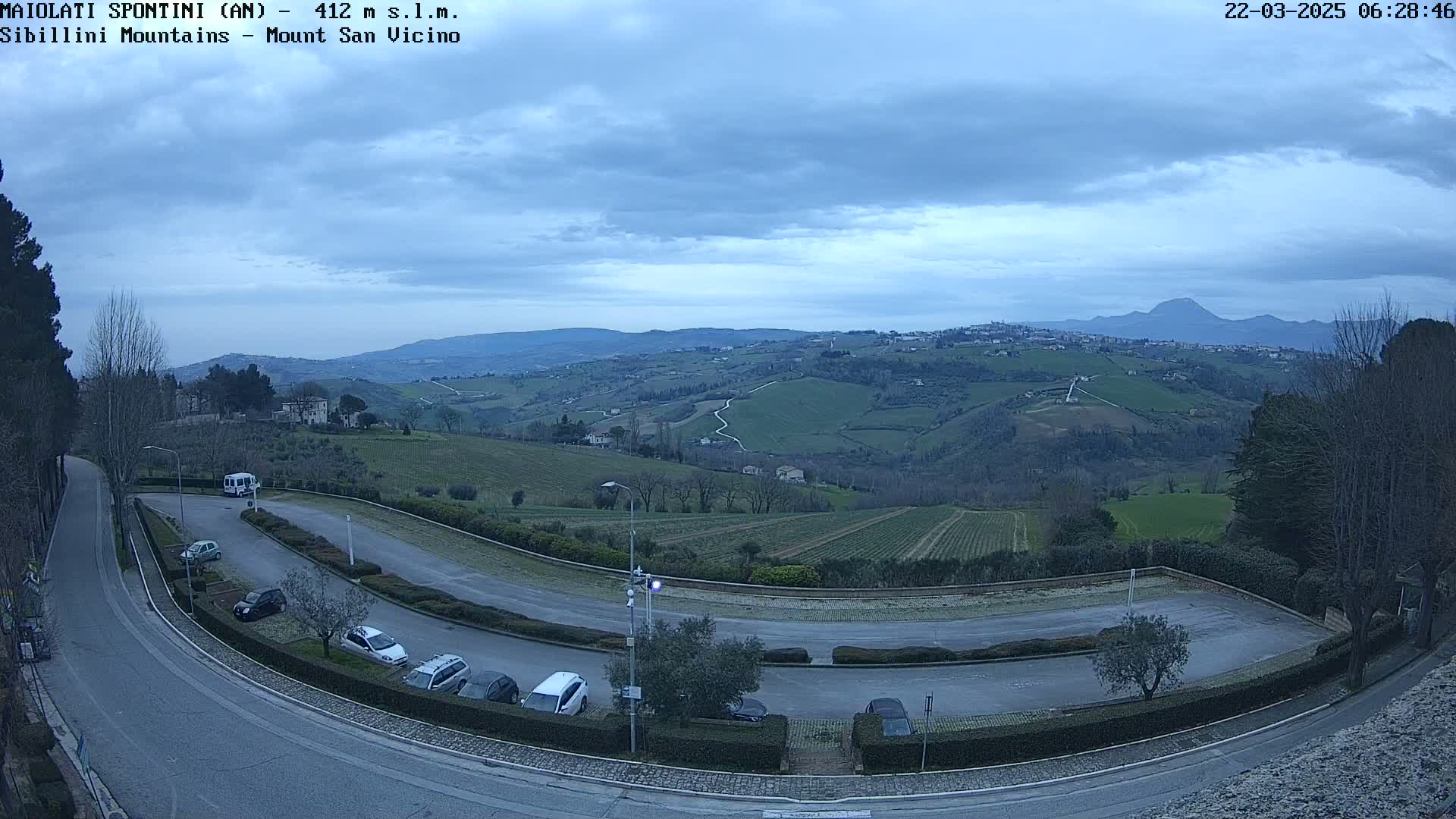 A winding road curves through a parking lot overlooking a hilly landscape under a cloudy sky.
