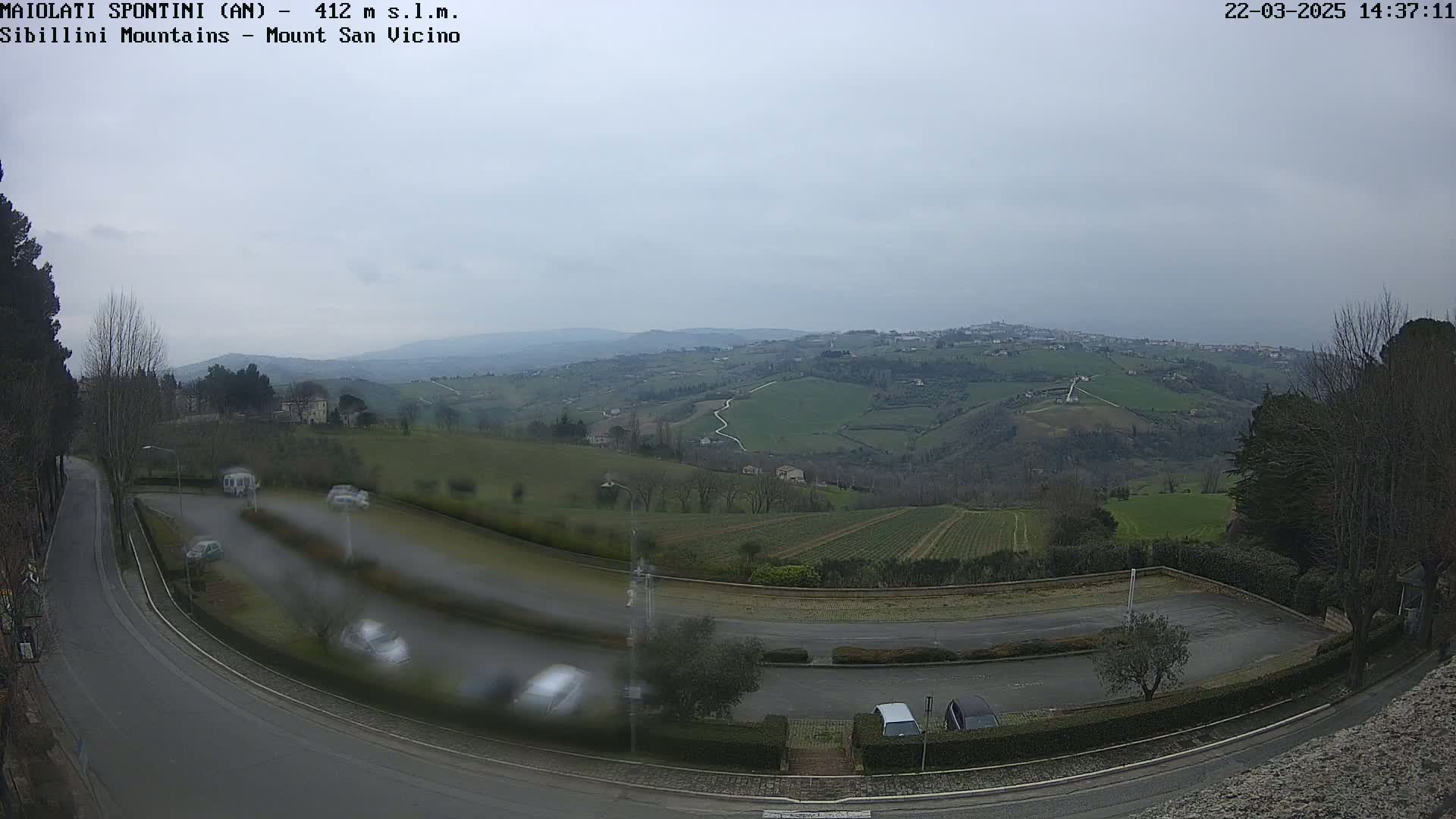 A parking lot overlooks a hilly landscape with a town in the distance under an overcast sky.