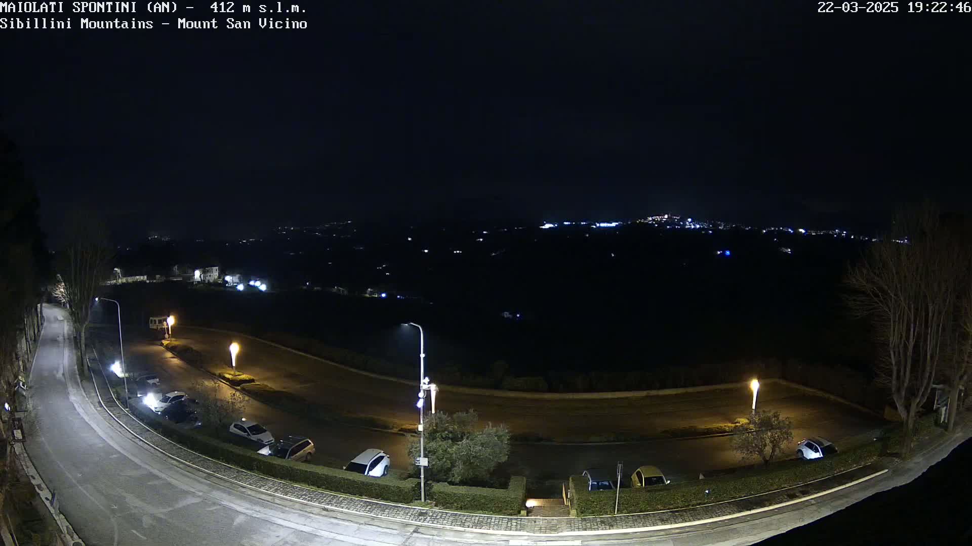 A nighttime, high-angle view shows a curving road with parked cars and illuminated streetlights, overlooking a dark, distant town speckled with lights.