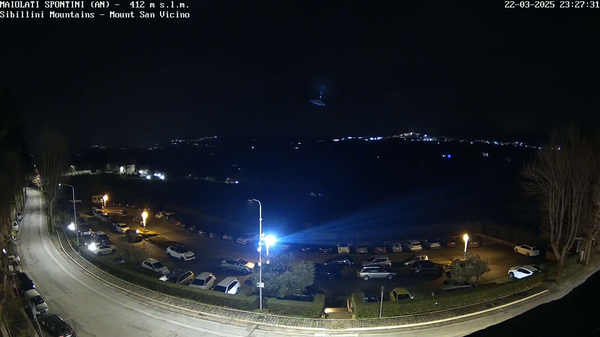 A nighttime aerial view of a parking lot with numerous cars, a curving road, and a distant town illuminated under a clear, dark sky.
