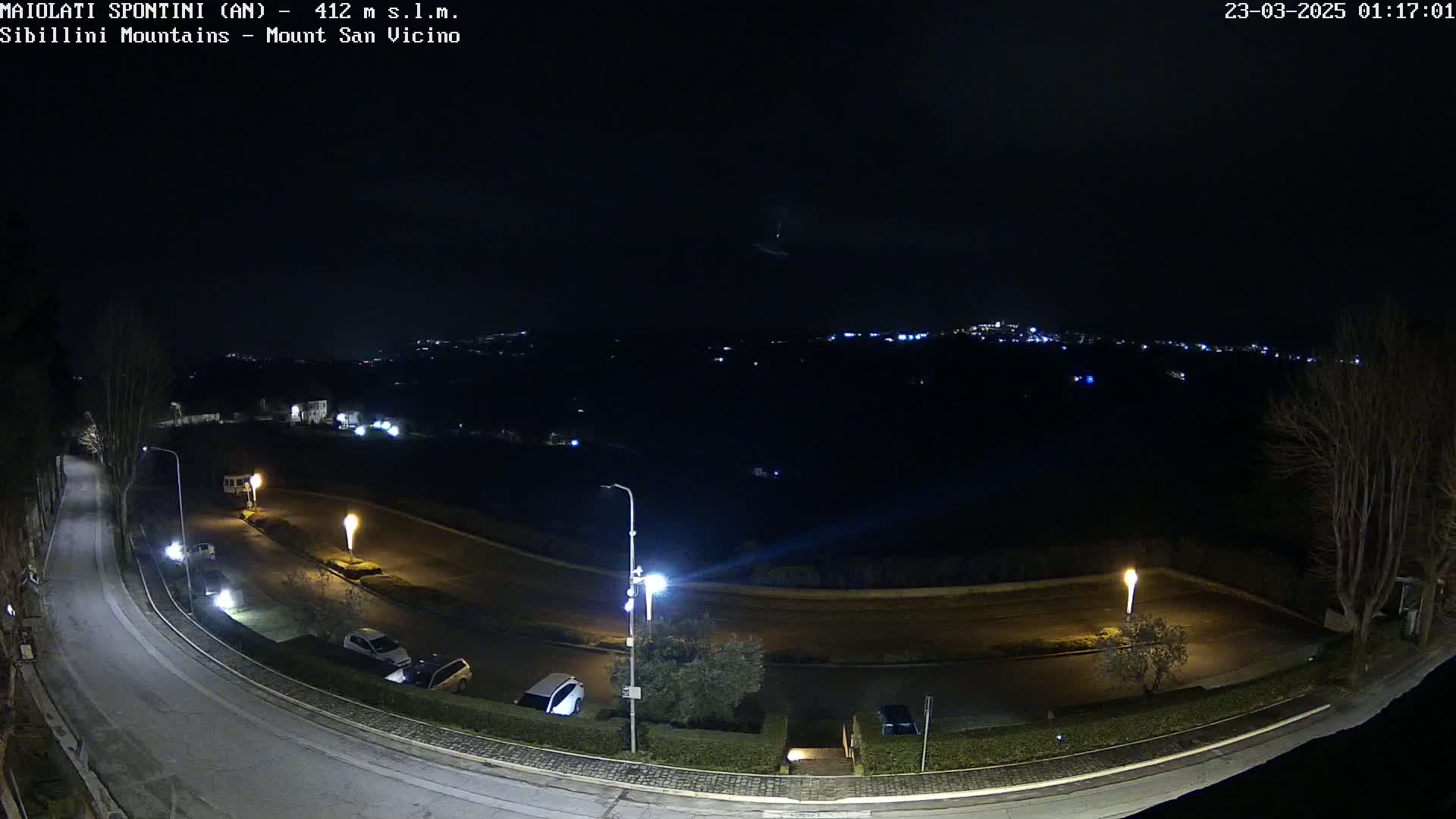 A nighttime, high-angle view of a curved road with parked cars and streetlights, overlooking a dimly lit town nestled in a valley.