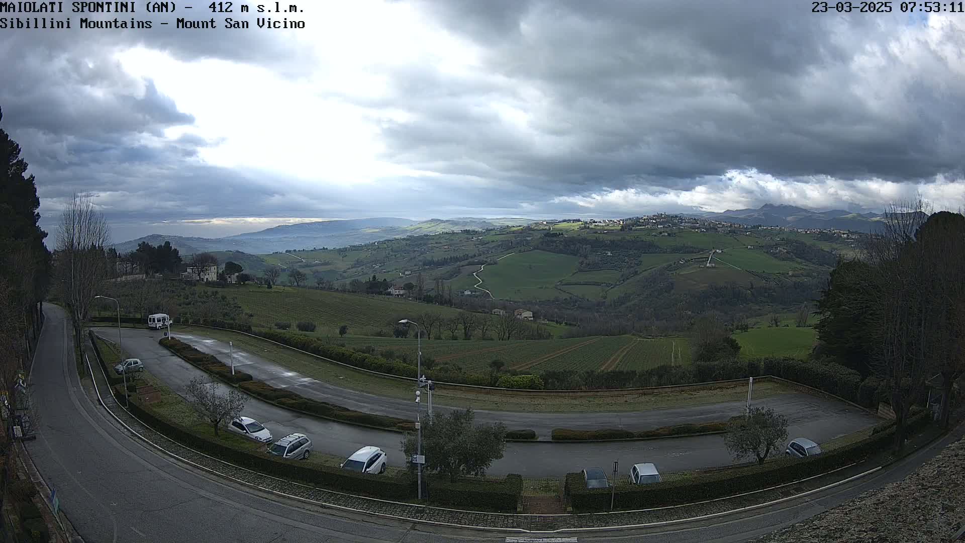 A mostly cloudy sky overlooks a hilly landscape with a parking lot and road curving through it, showing farmland and a village in the distance.