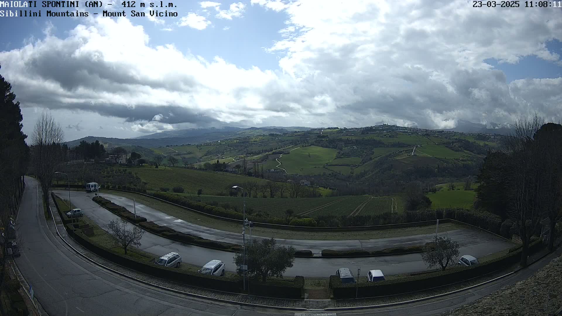 A wet road curves through a parking lot overlooking a hilly landscape of green fields and dark trees under a partly cloudy sky.