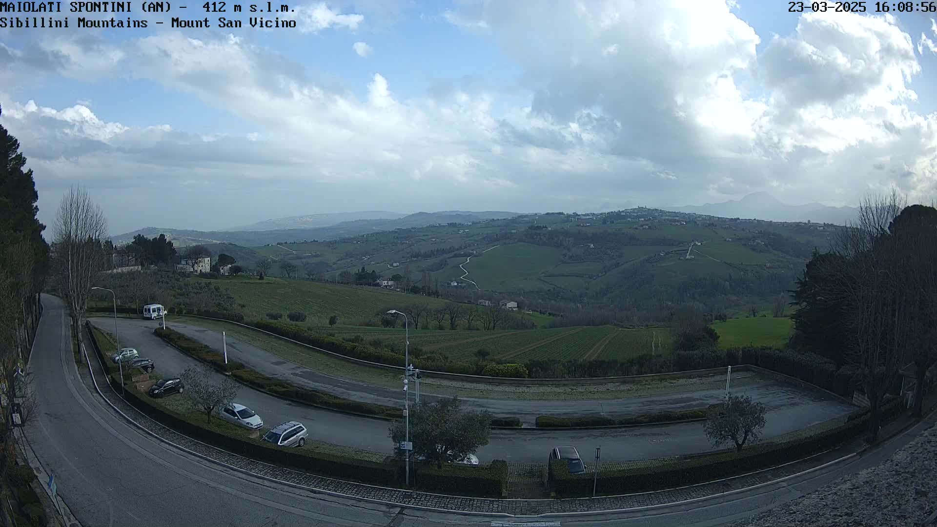 A mostly cloudy day reveals a view of rolling green hills and a small parking lot with several cars, overlooked by a curving road.