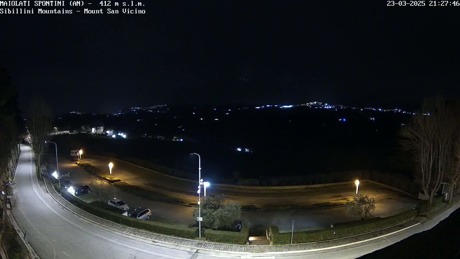 A nighttime, high-angle view of a paved road curving through a parking lot and sparsely lit landscape, with a distant city illuminated in the background.
