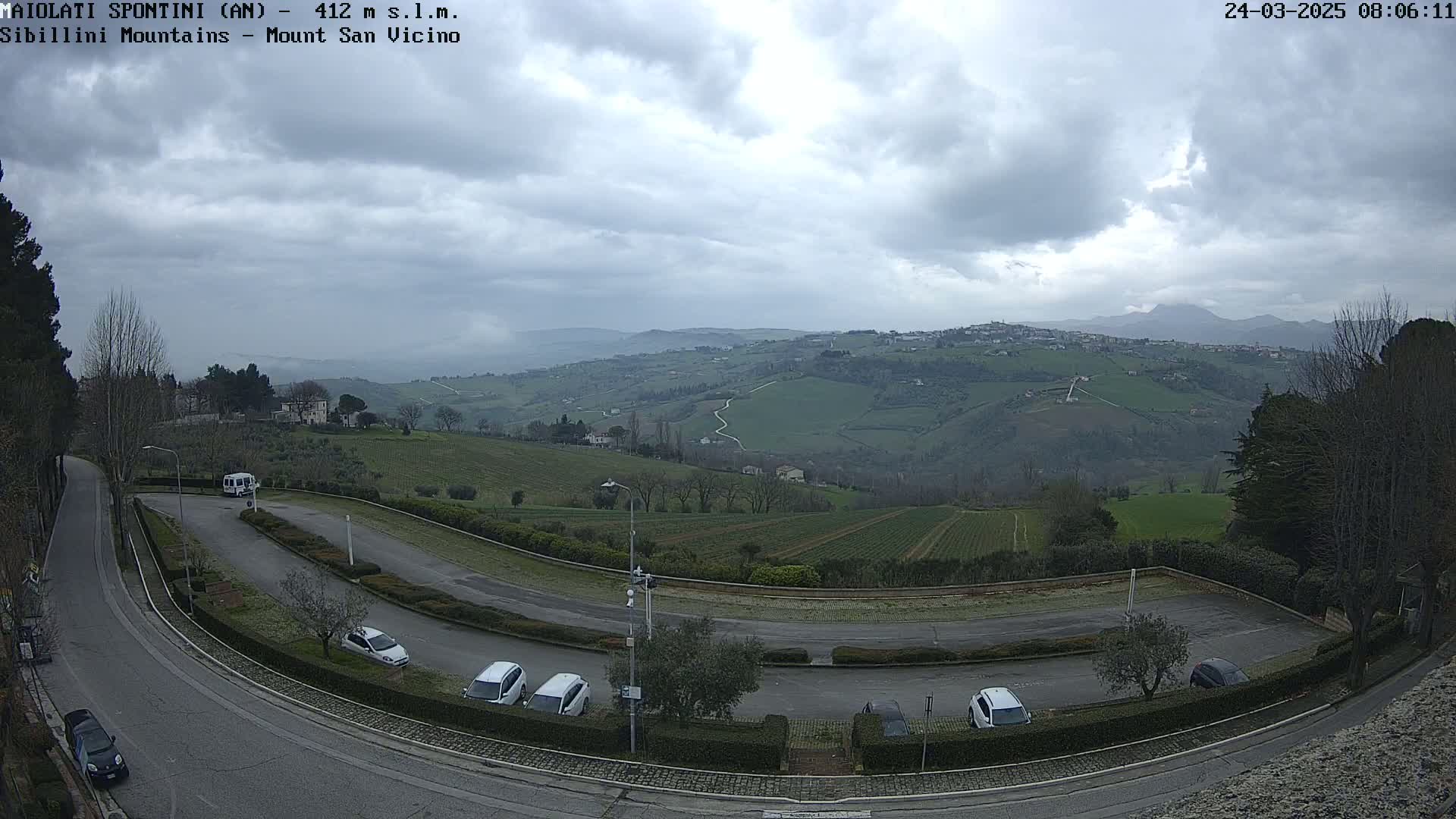 A parking lot with several cars is situated on a hill overlooking a valley of rolling green fields under a cloudy sky.