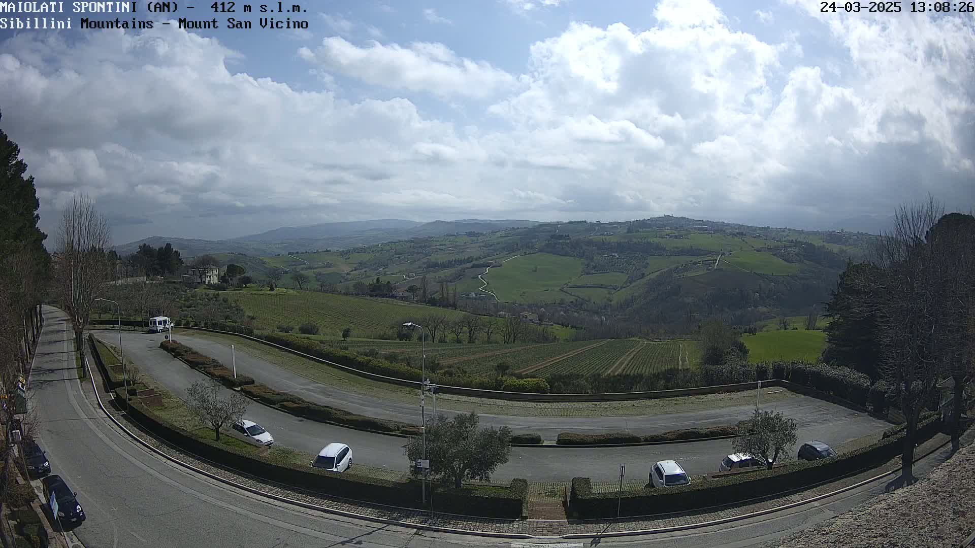 A parking lot with a few cars curves around a hillside overlooking a vast, rolling green landscape under a partly cloudy sky.