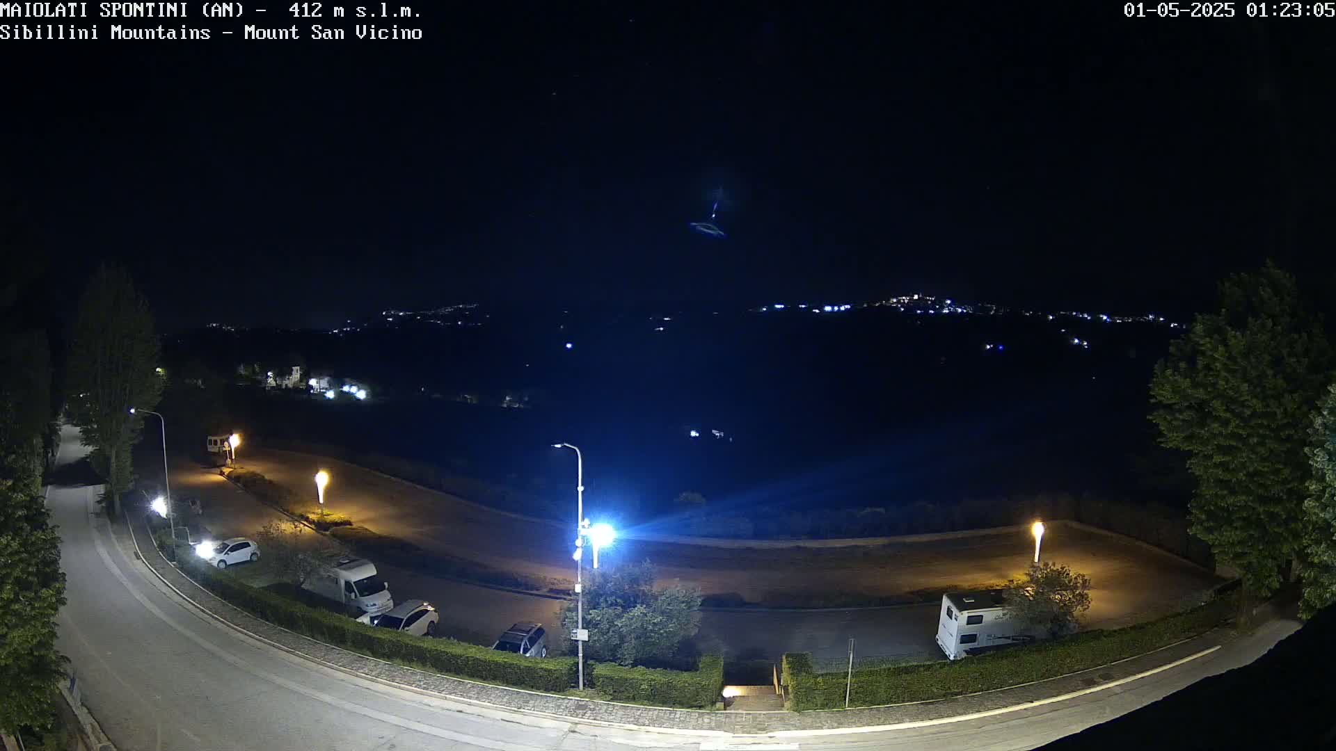 A nighttime view of a parking lot with several vehicles, illuminated by streetlights, overlooking a distant town under a clear, dark sky.