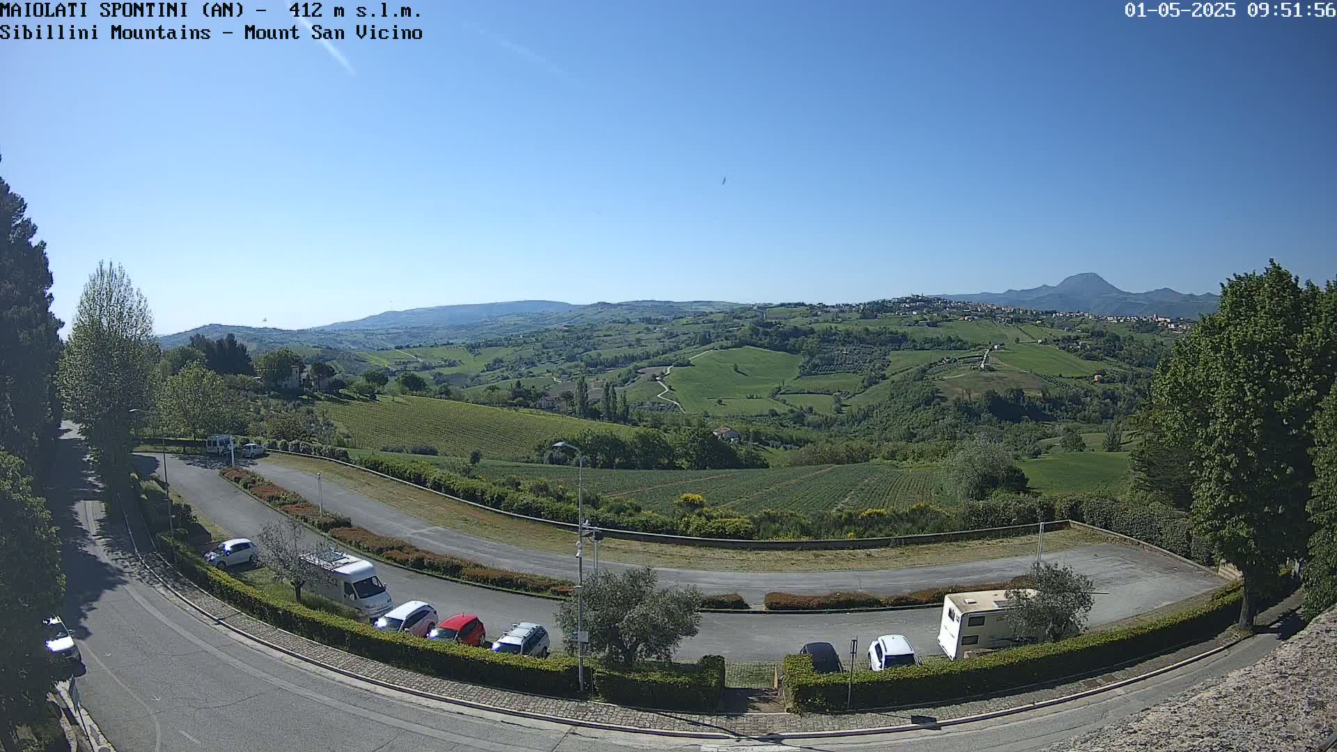 A paved parking lot with several cars and camper vans overlooks a sunny, verdant valley dotted with vineyards and distant hills under a clear blue sky.