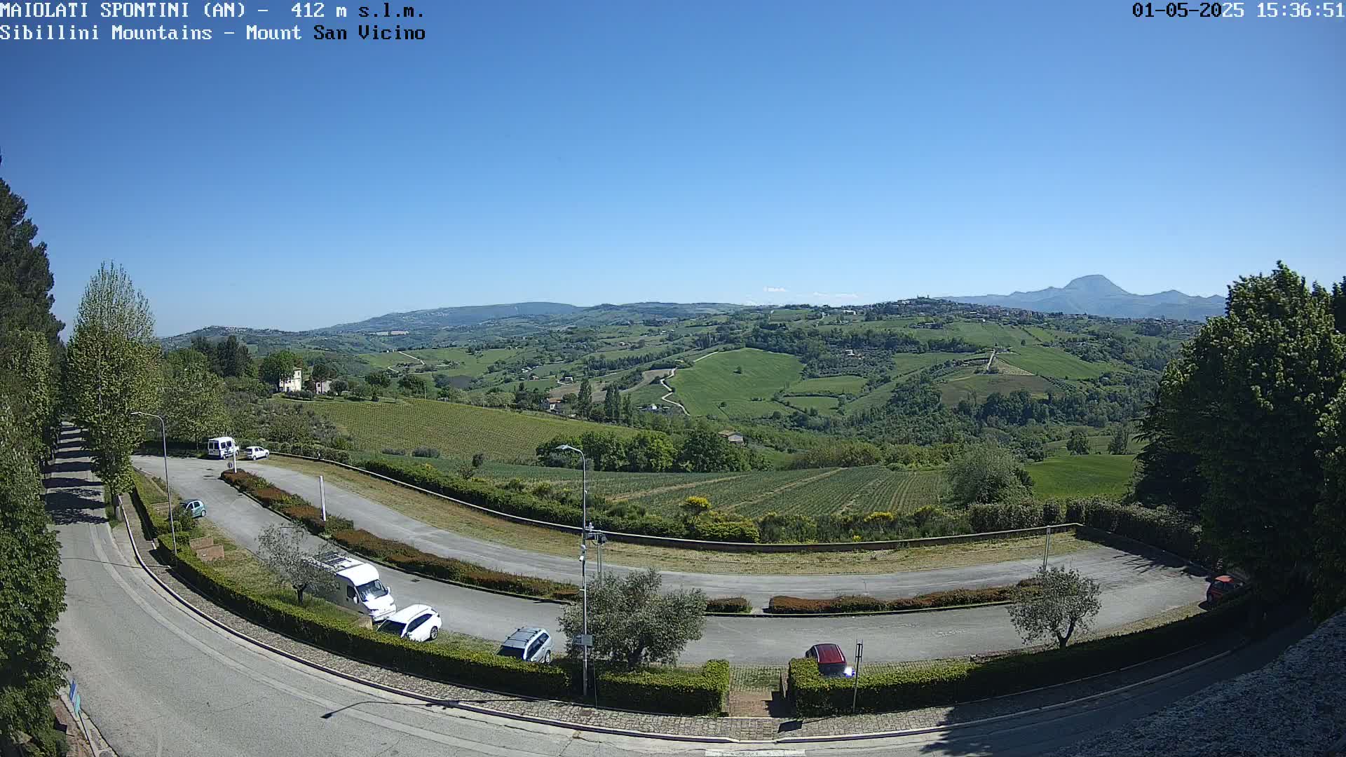 A sunny day reveals a winding road curving through a parking lot, overlooking a vast expanse of rolling green hills and vineyards under a clear blue sky, with a distant mountain range visible on the horizon.