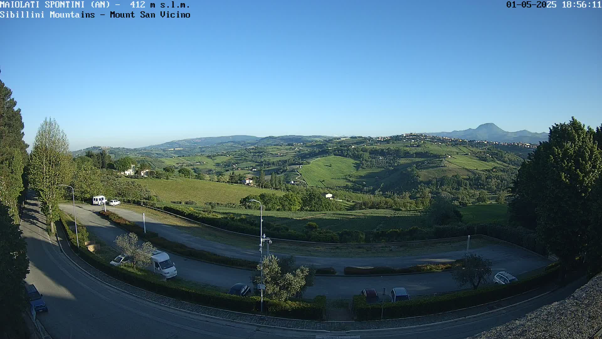 A panoramic view of a hilly landscape under a clear blue sky shows a parking lot and road curving through the foreground, with vineyards and distant mountains in the background.