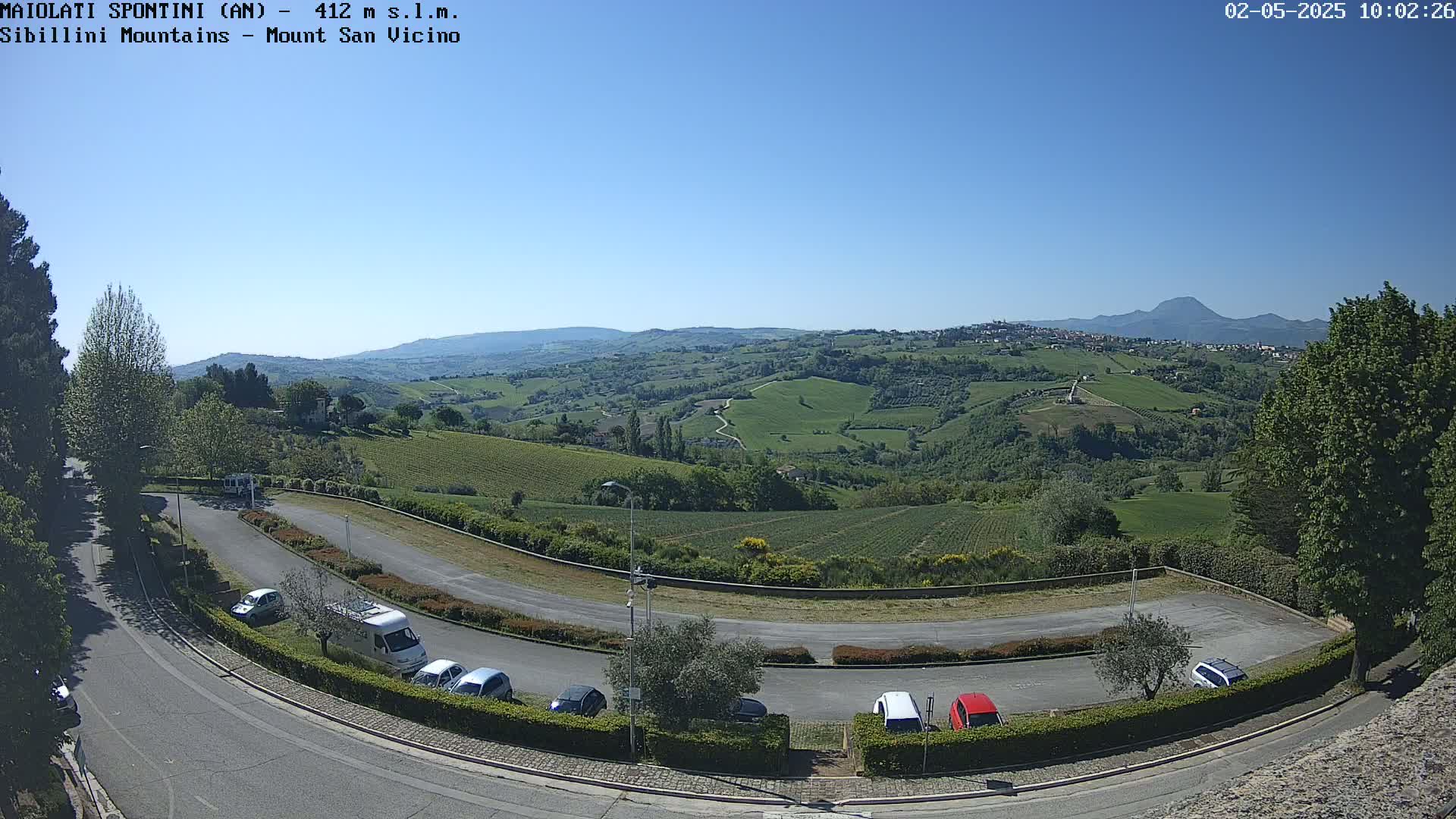 A clear, sunny day reveals a panoramic view of rolling green hills, vineyards, and a parking lot with several cars, from a high vantage point.