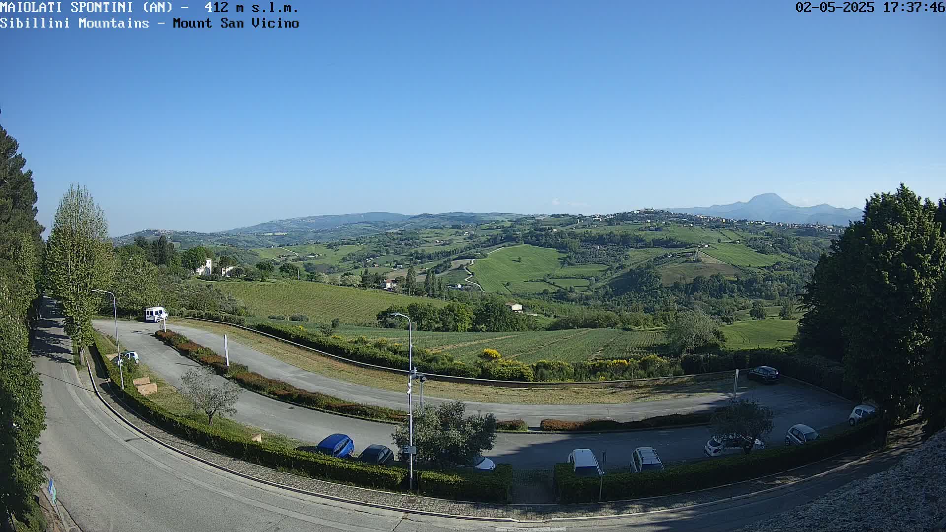 A winding road curves through a parking lot overlooking a sunny, verdant valley dotted with buildings and farmland, mountains visible in the distance under a clear blue sky.