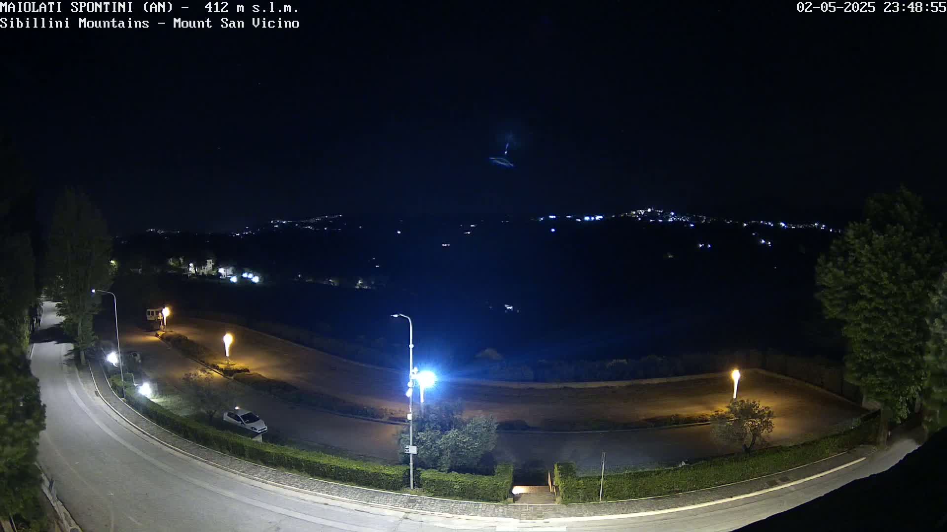 A nighttime, overhead view of a winding road with parked cars and illuminated streetlights, overlooking a distant, dimly lit town under a dark sky with a visible streak of light.