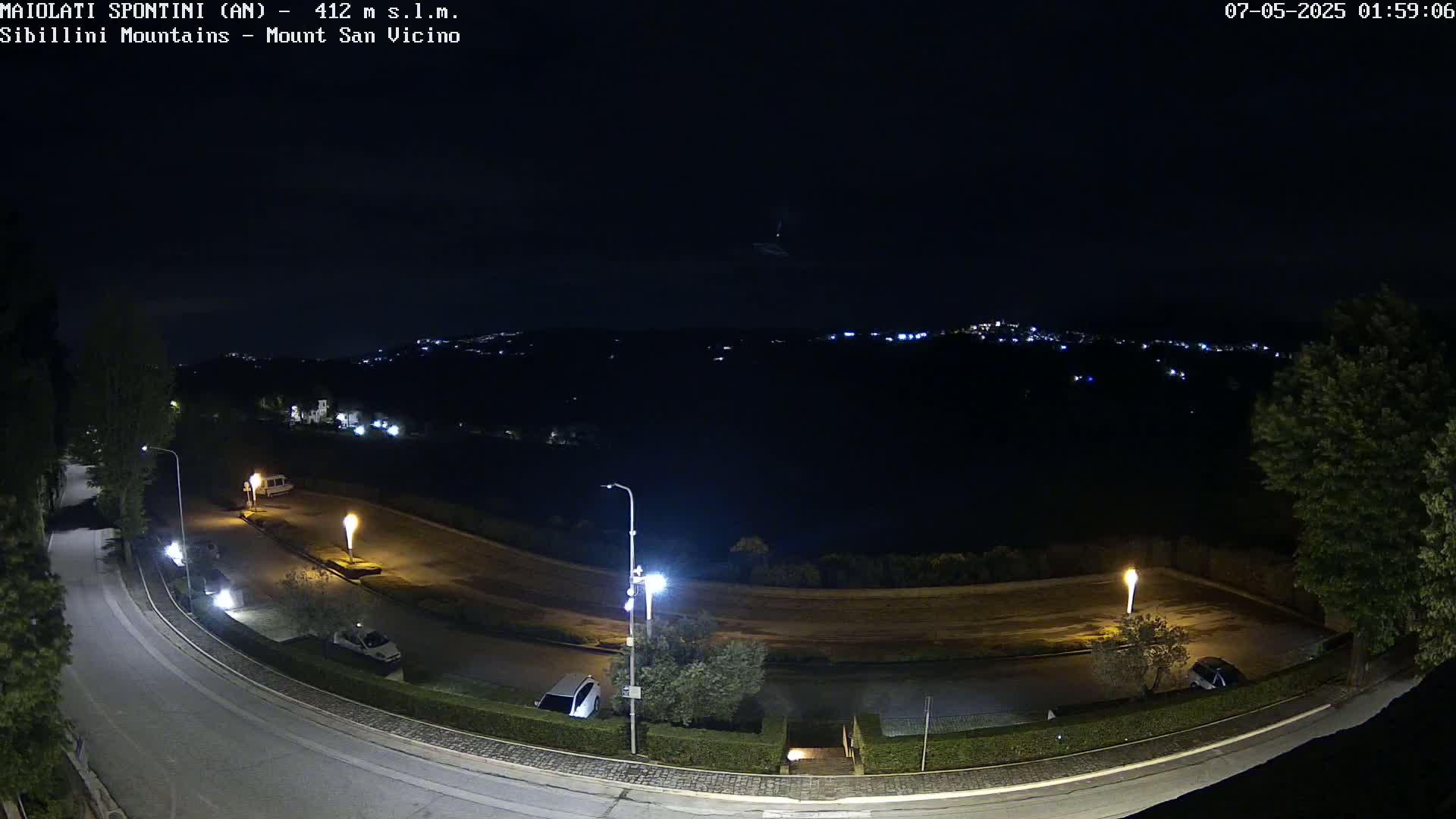 A nighttime, high-angle view shows a mostly empty parking lot and roadway curving to the left, illuminated by streetlights, with a distant, dimly lit town visible in the background under a dark, clear sky.