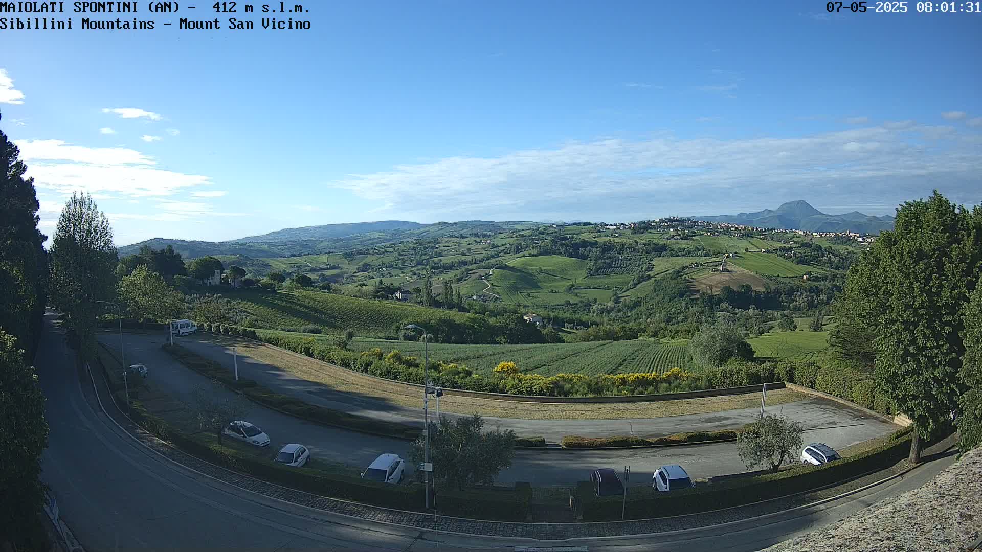 A panoramic view of a hilly landscape with vineyards and fields under a mostly clear, sunny sky, showing a parking lot and road in the foreground.