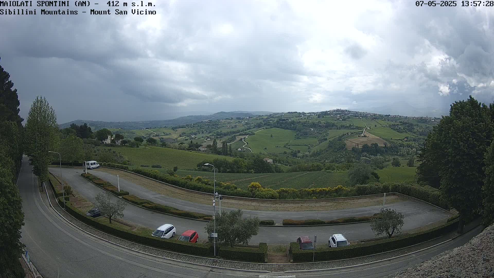 A winding road curves through a parking lot overlooking a hilly landscape of green vineyards under a partly cloudy sky.