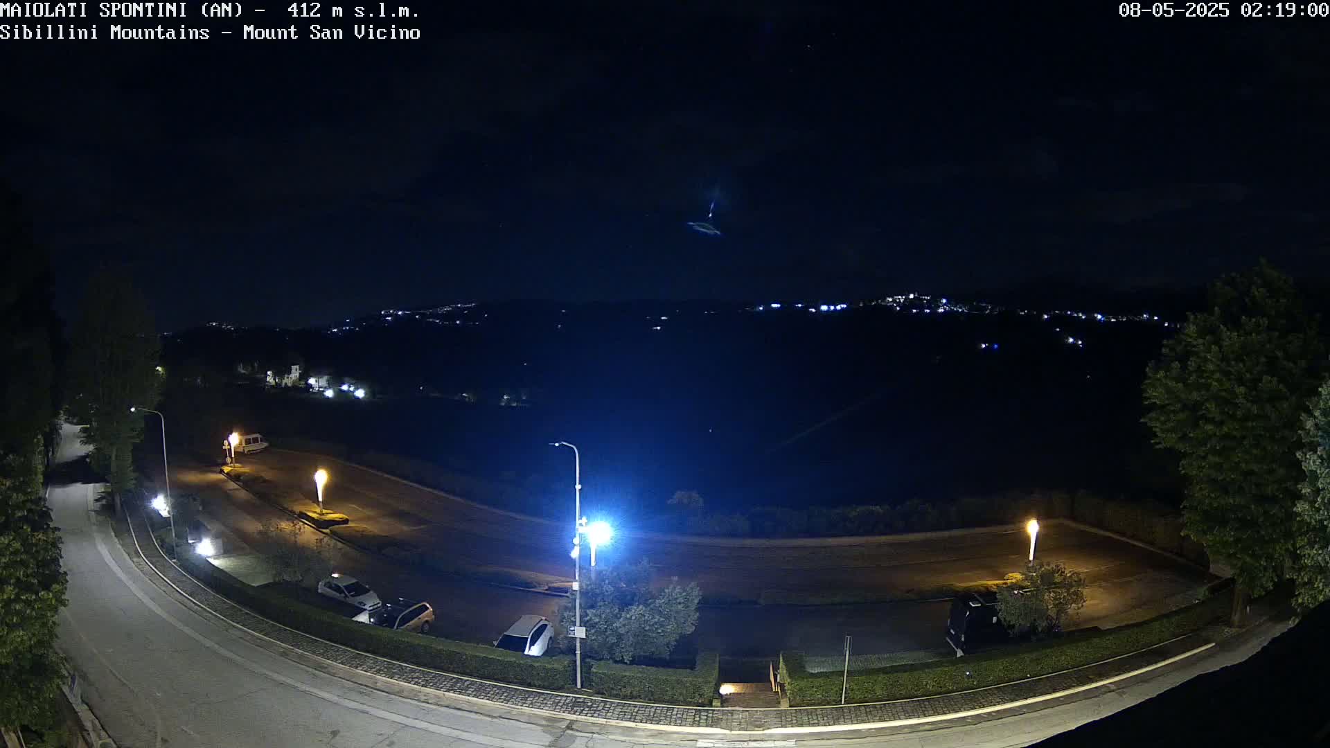 A nighttime view of a curving road with parked cars, illuminated by streetlights, overlooking a distant town under a dark, clear sky showing a meteor.