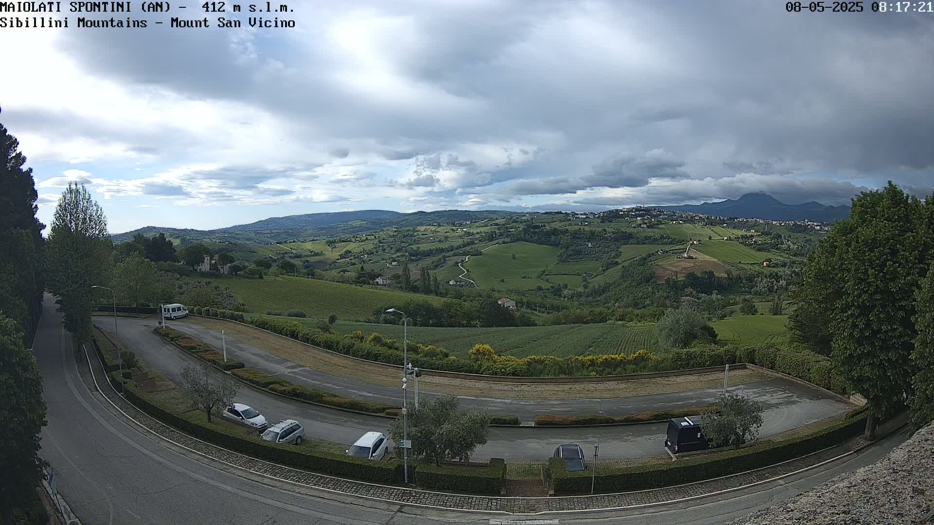 A parking lot with several cars overlooks a hilly landscape of green fields and vineyards under a partly cloudy sky.