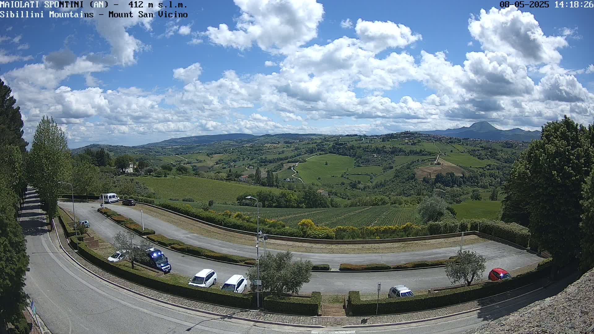A partly cloudy sky overlooks a hilly landscape of vineyards and fields, with a parking lot and road in the foreground.