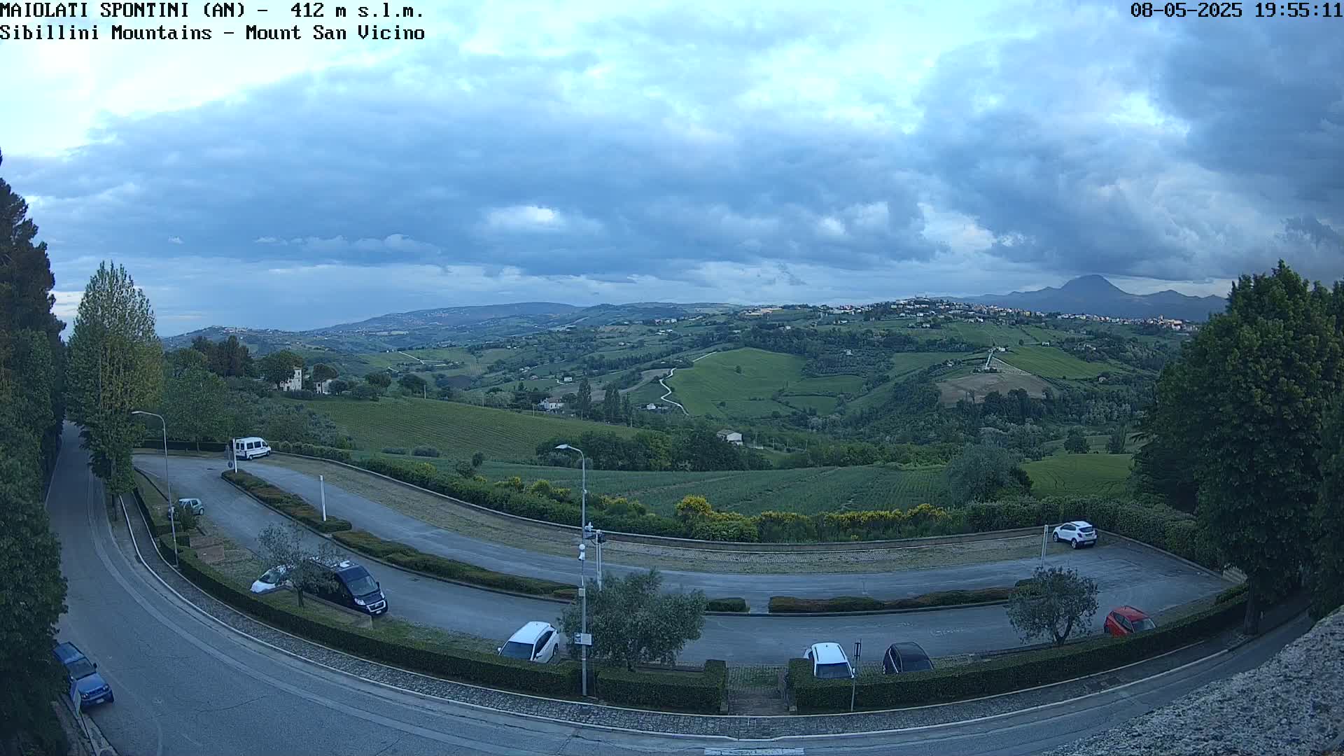 A curved road with parked cars overlooks a hilly landscape under a partly cloudy sky.