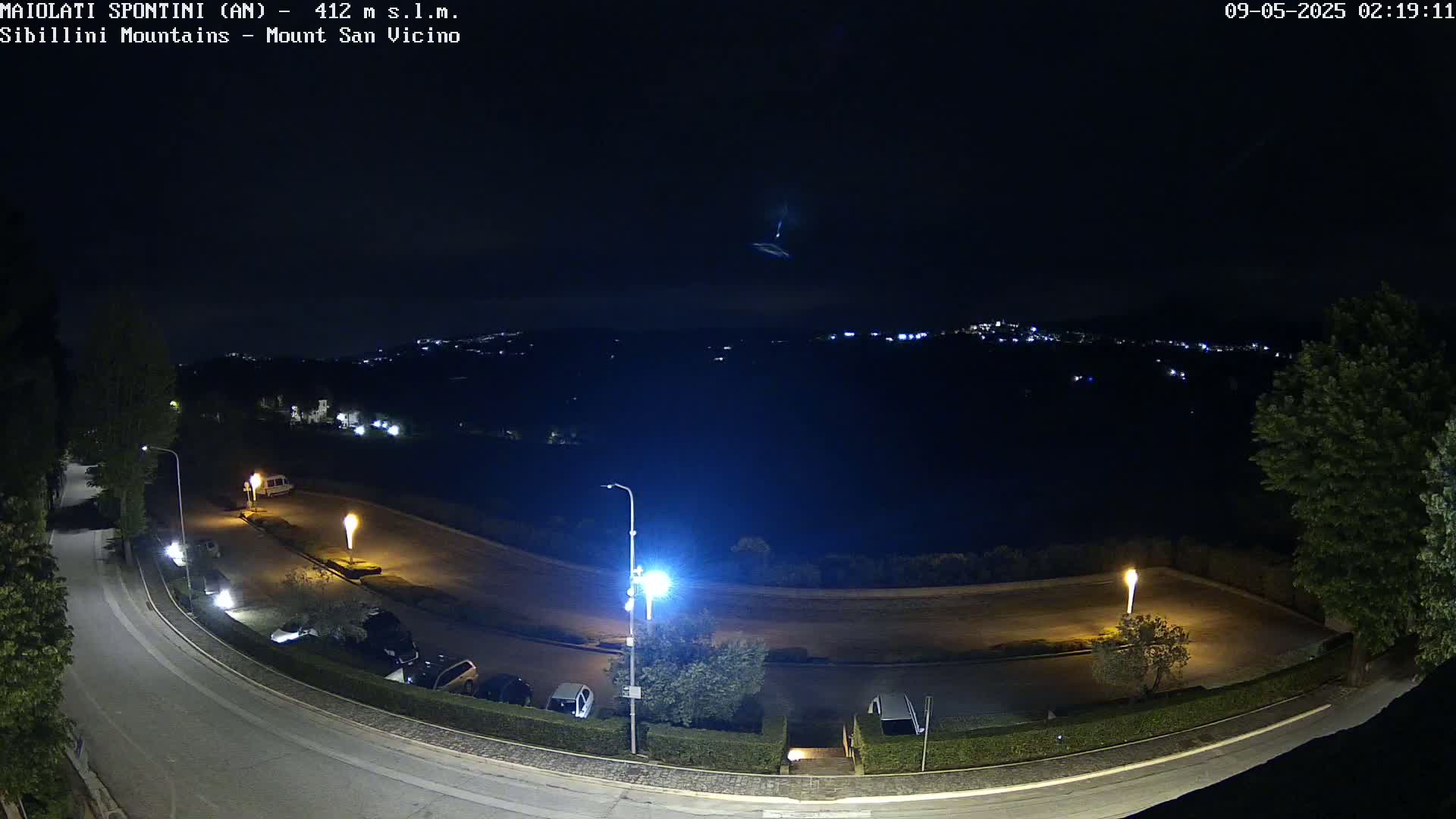 A nighttime, high-angle view of a curved road with parked cars and illuminated lights, overlooking a distant town nestled in a dark, mountainous landscape under a clear sky.