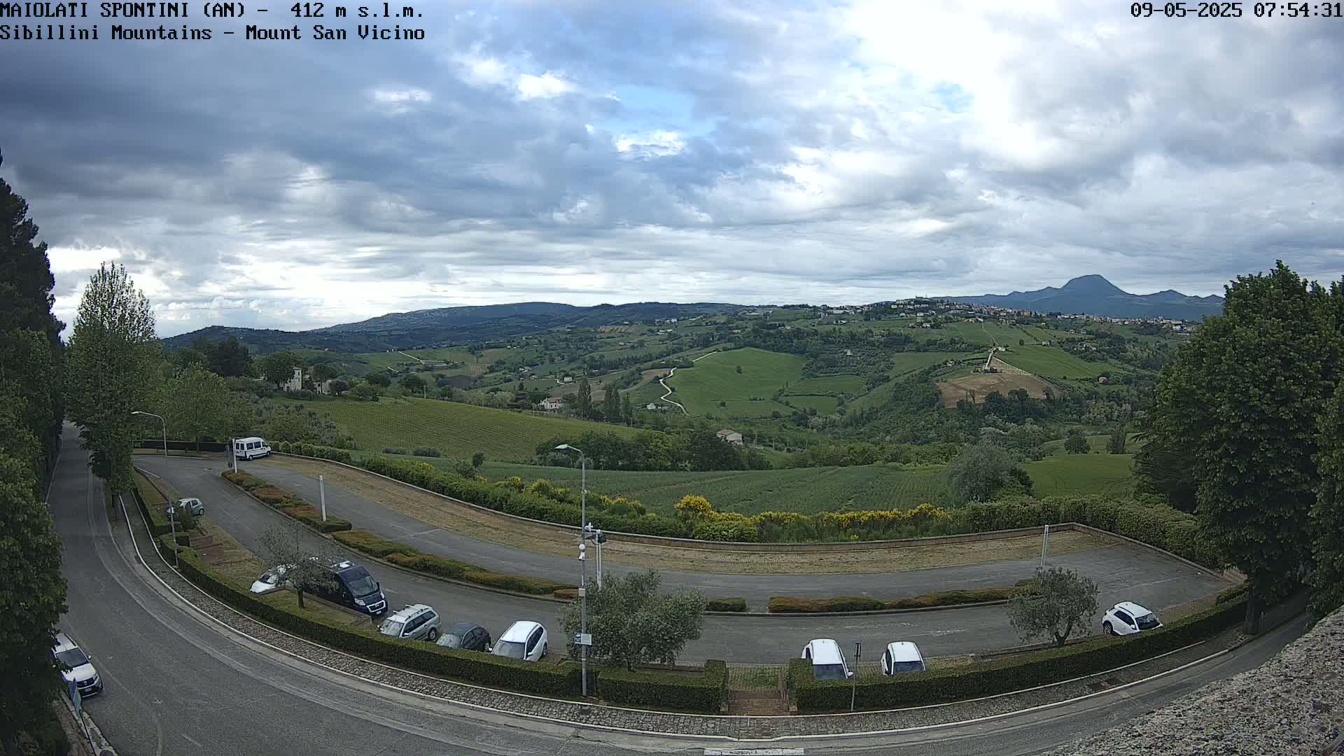 A mostly cloudy day overlooks a paved parking lot and curving road, nestled amongst verdant rolling hills and farmland, with a distant mountain range visible in the background.