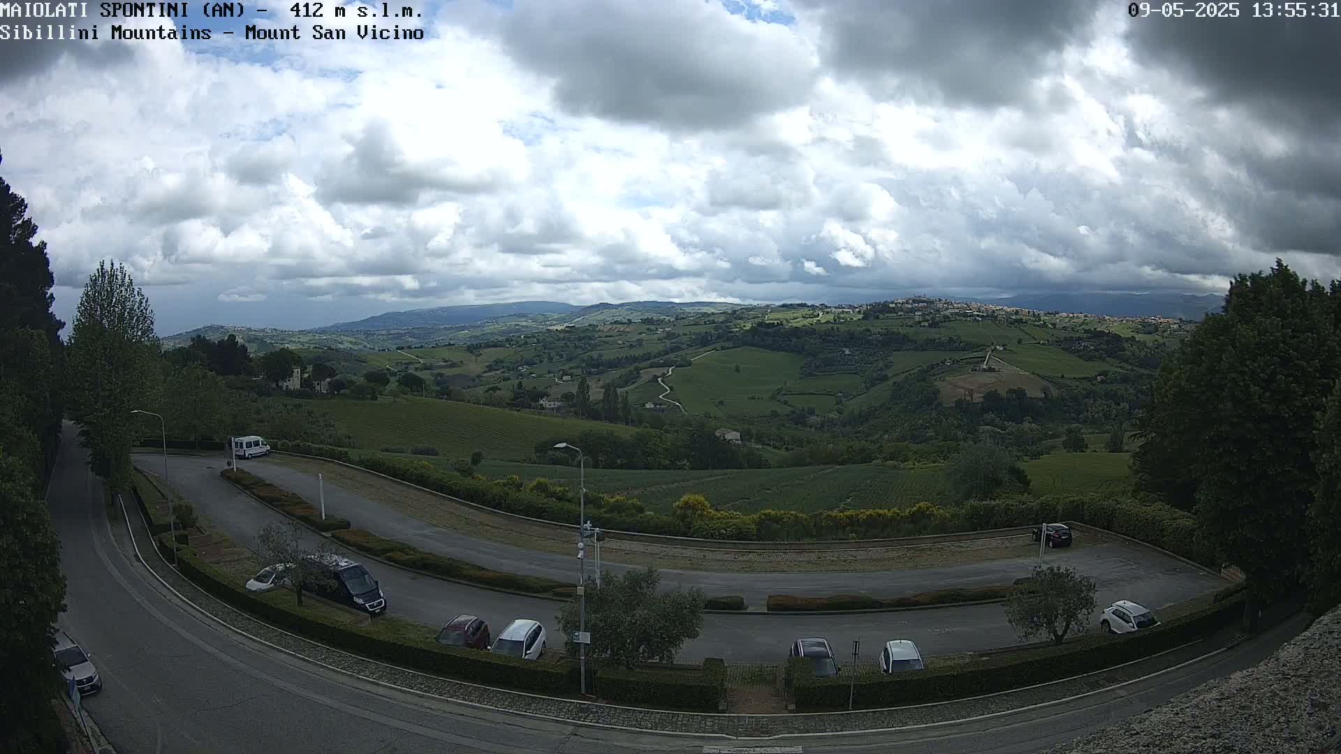 A mostly cloudy sky overlooks a winding road and parking lot nestled in a hilly, verdant landscape.