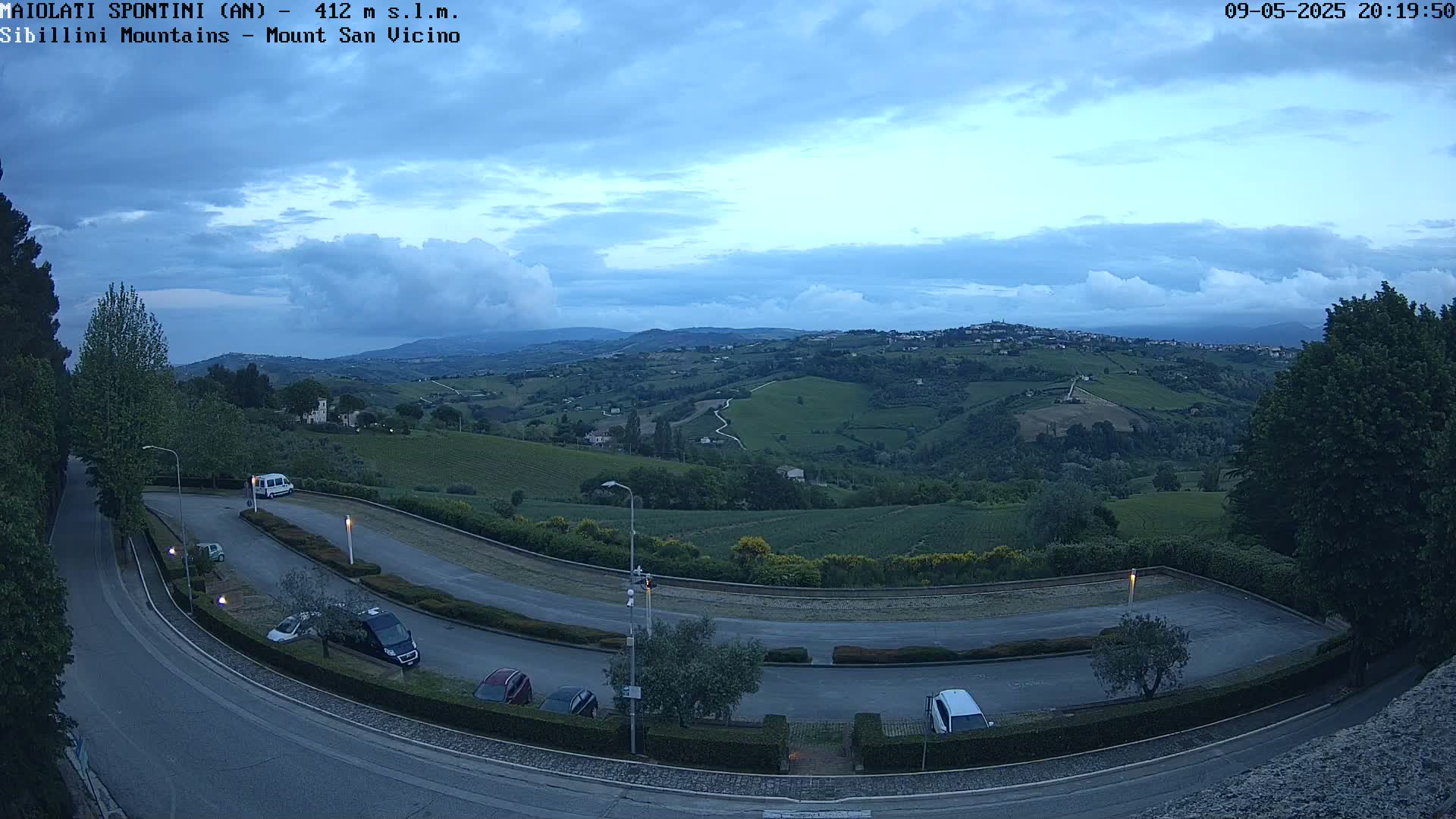 A mostly cloudy twilight sky overlooks a hilly landscape with vineyards and a parking lot containing several cars and a van.