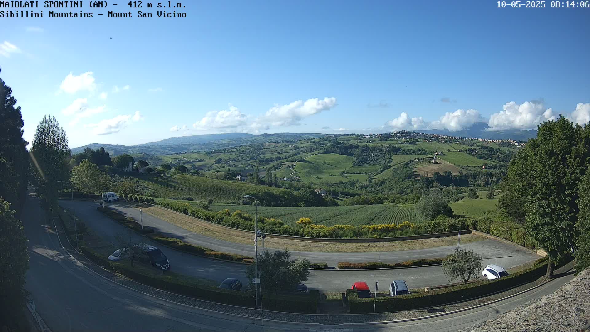 A mostly clear, sunny day reveals a panoramic view of rolling green hills, a parking lot with several cars, and a winding road curving through the landscape.