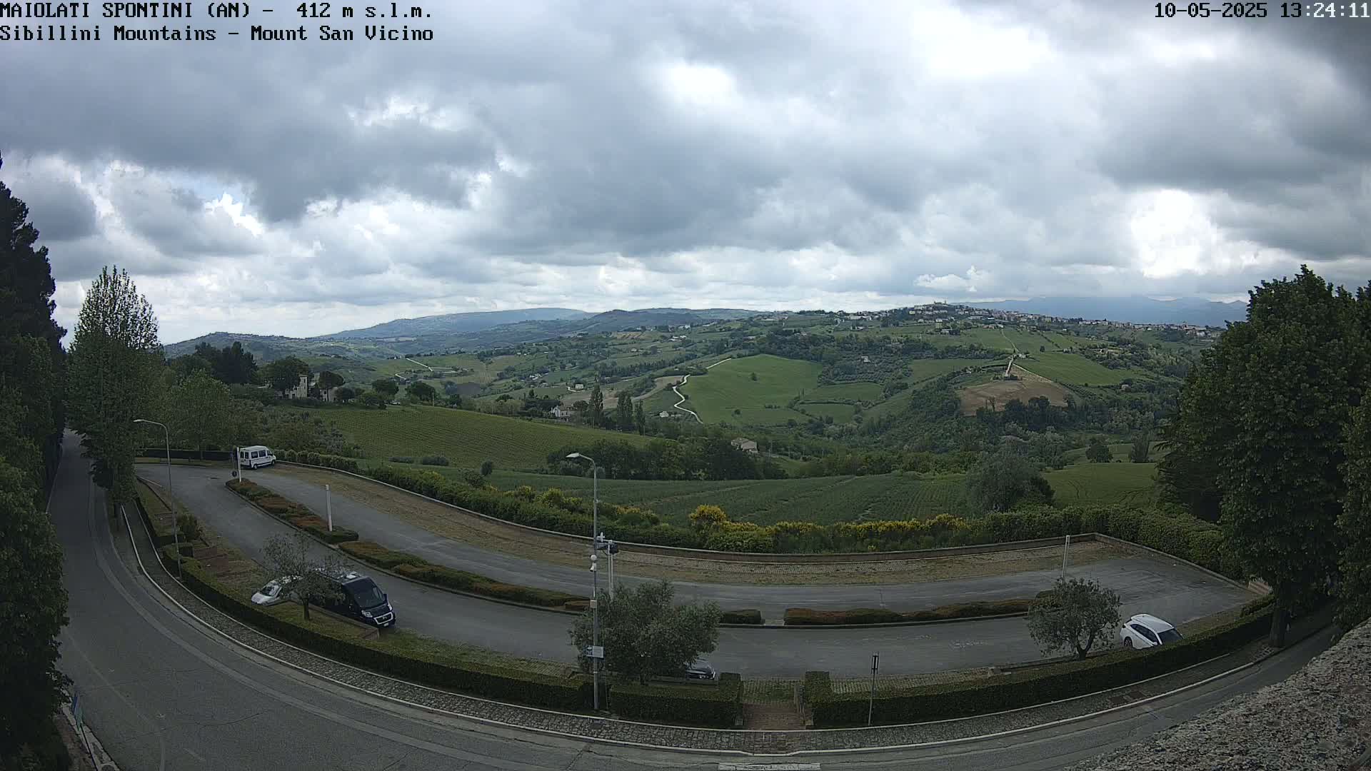 A winding road curves through a parking lot overlooking a hilly landscape of green fields and vineyards under a cloudy sky.