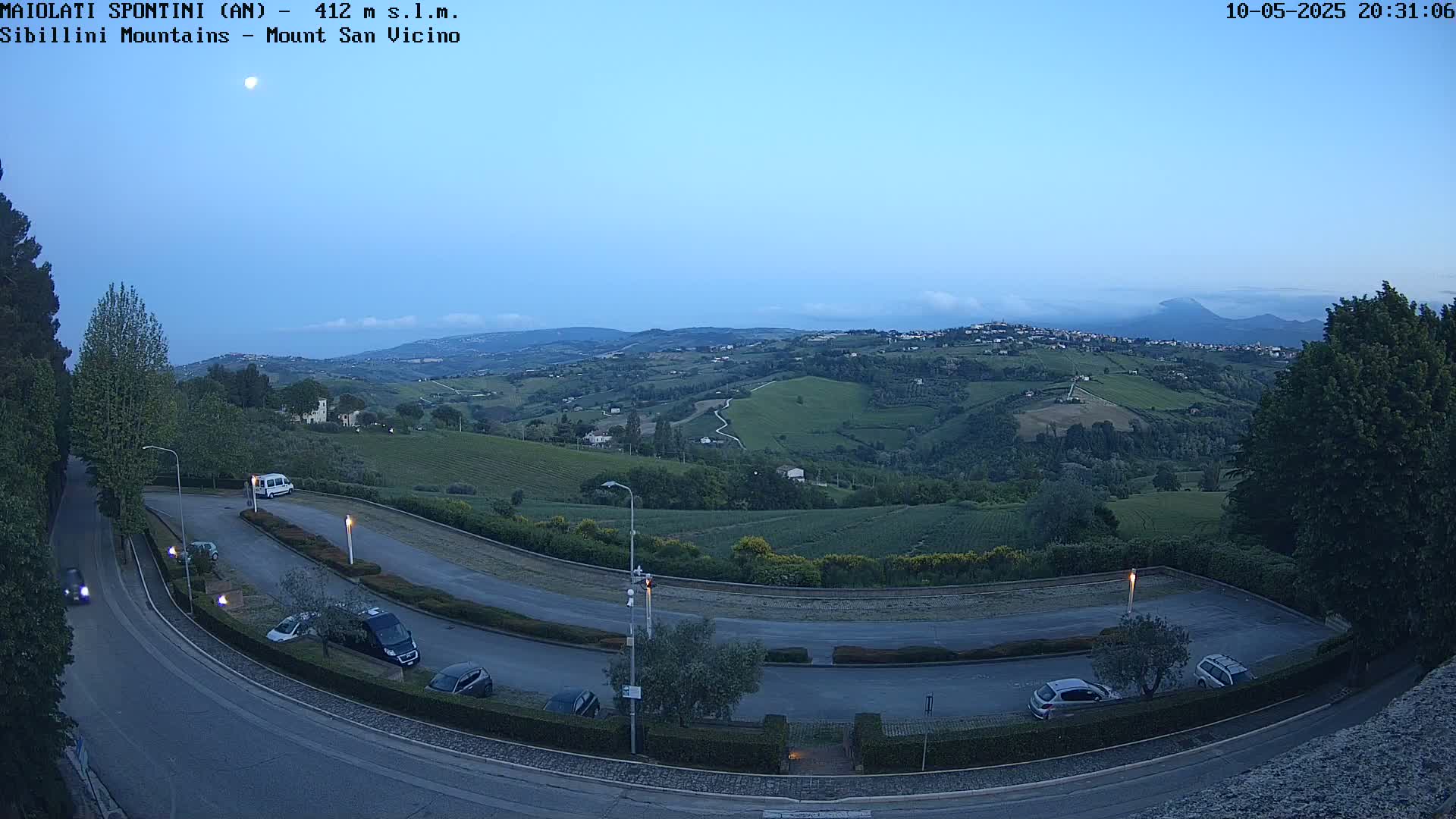 A moonlit, dusk view of a hilly landscape with vineyards and a parking lot, showing several cars parked and a few driving on roads.