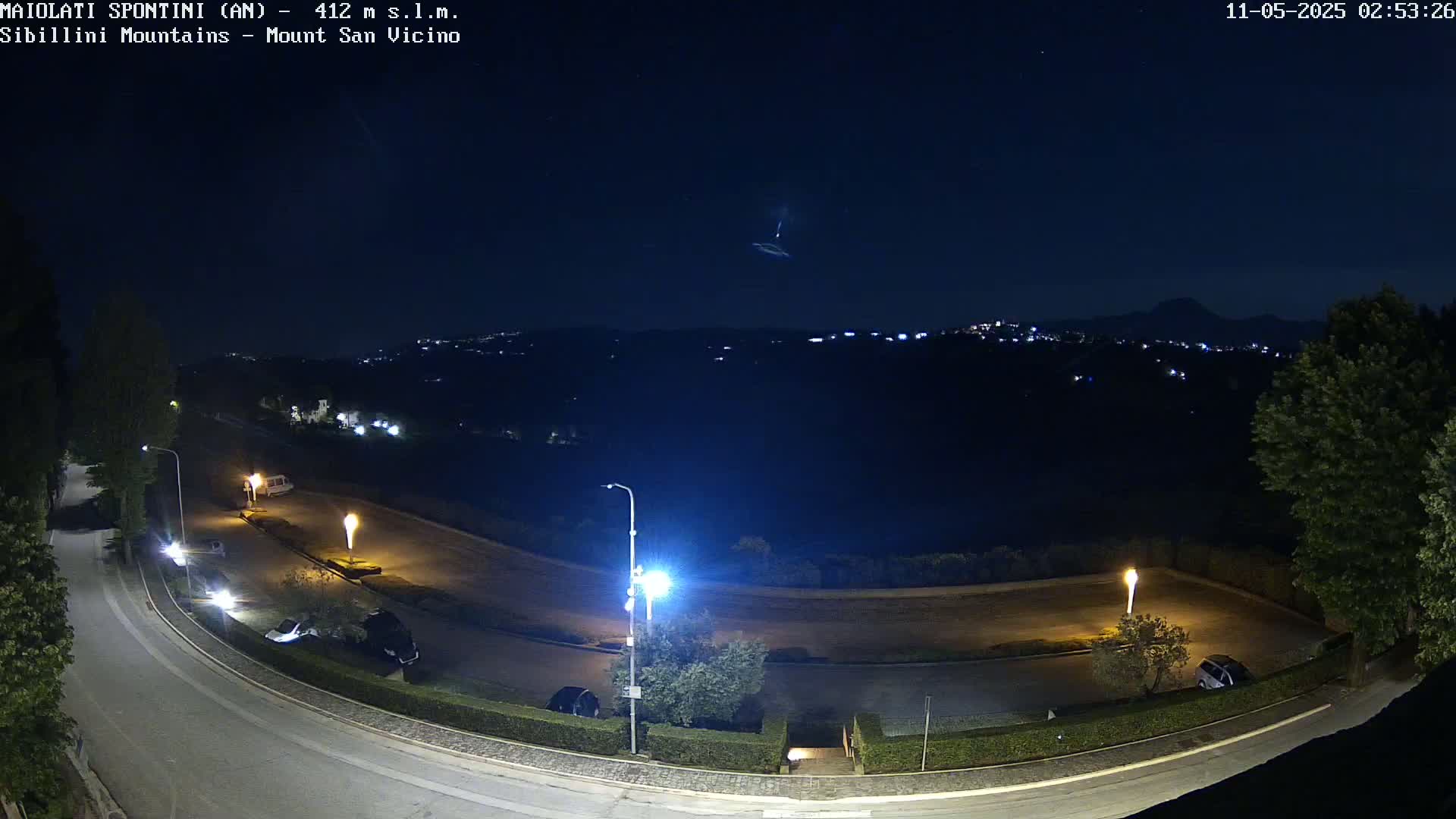 A nighttime, high-angle view shows a mostly empty parking lot and curving road illuminated by streetlights, overlooking a distant city nestled in dark hills under a clear, starlit sky.