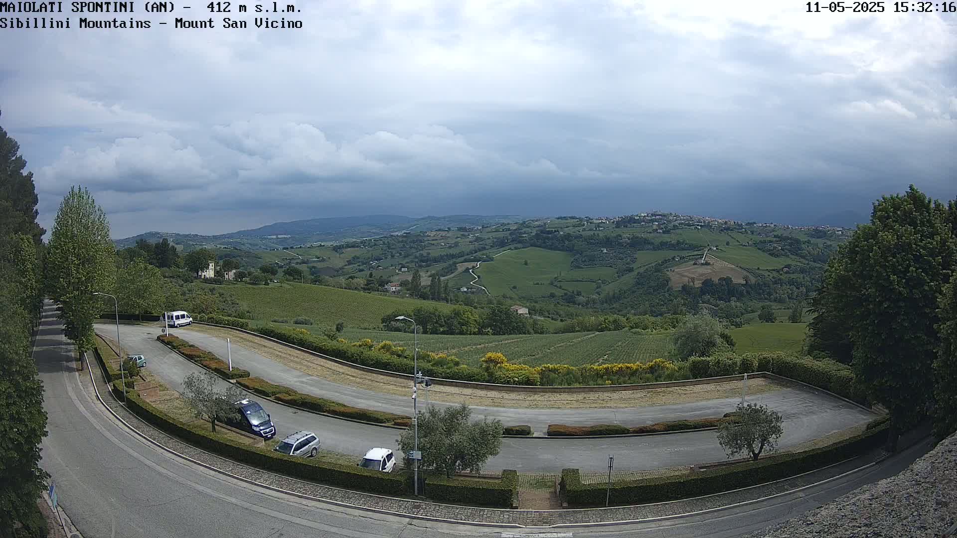 A paved road curves through a parking lot overlooking a hilly landscape of vineyards and trees under a mostly cloudy sky.