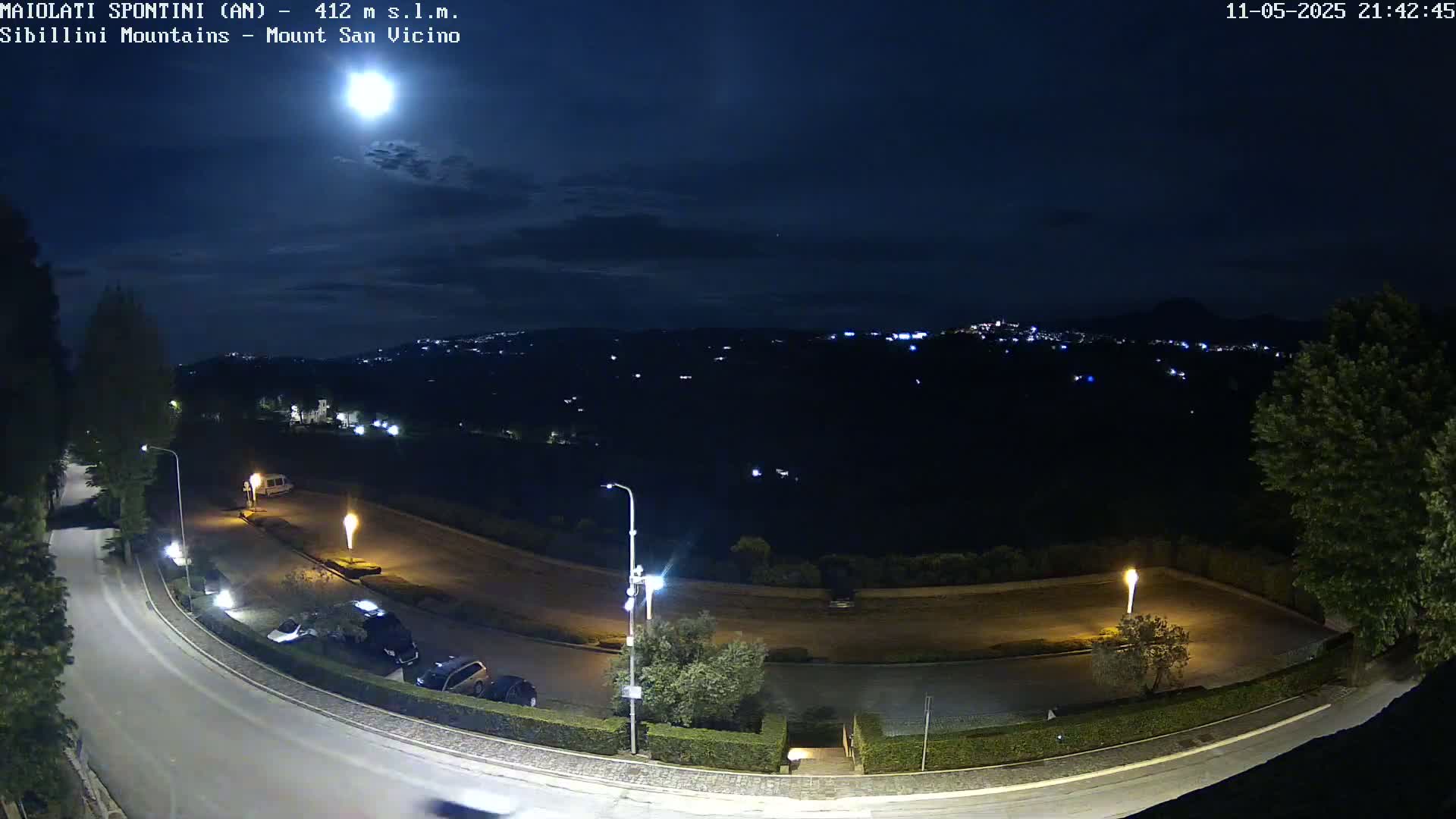 A nighttime, moonlit view of a curving road and parking lot, with distant city lights visible across a dark landscape.