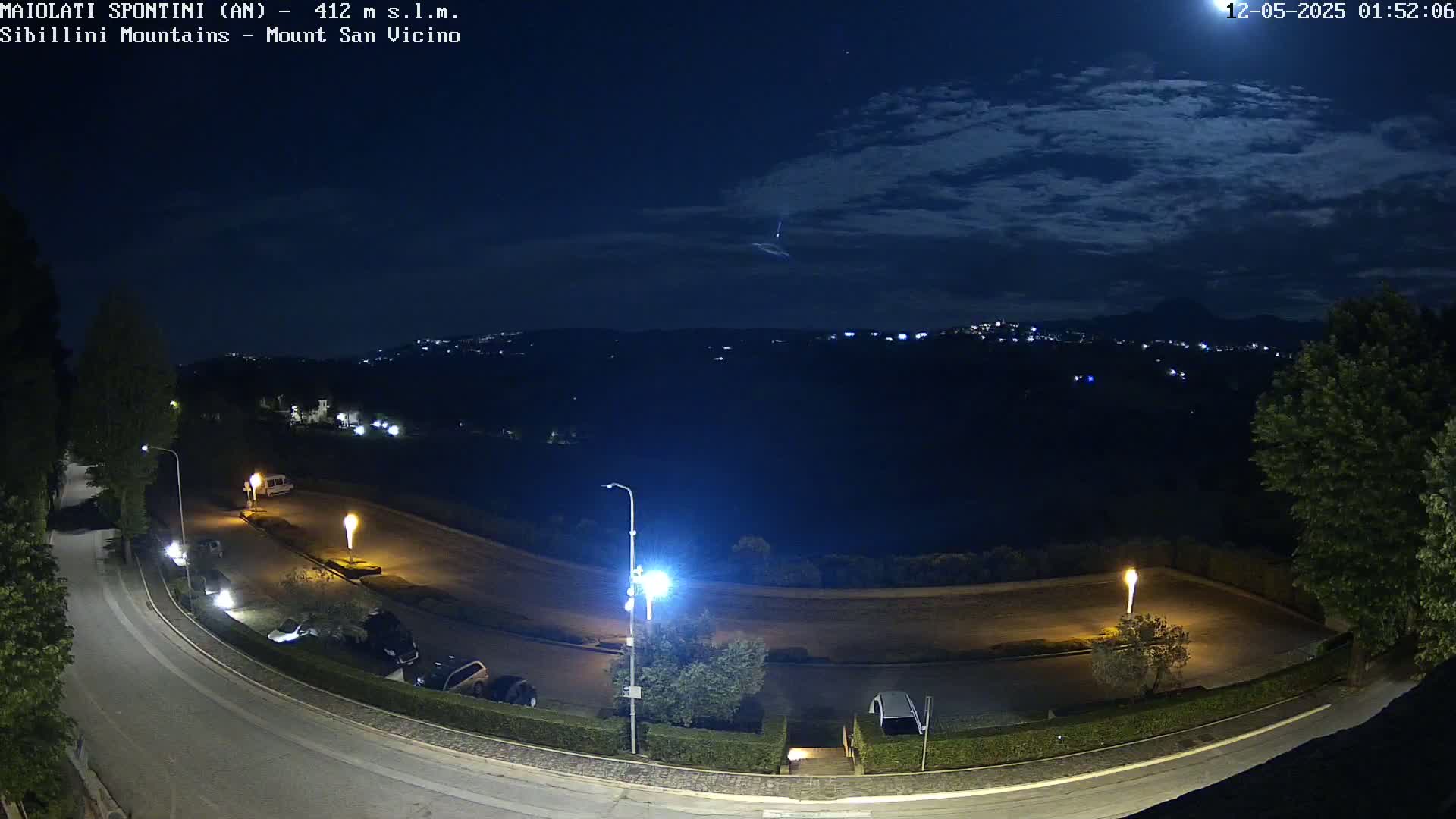 A nighttime view of a parking lot and curving road, illuminated by streetlights, with a distant, dimly lit town and a partly cloudy night sky showing a bright streak.