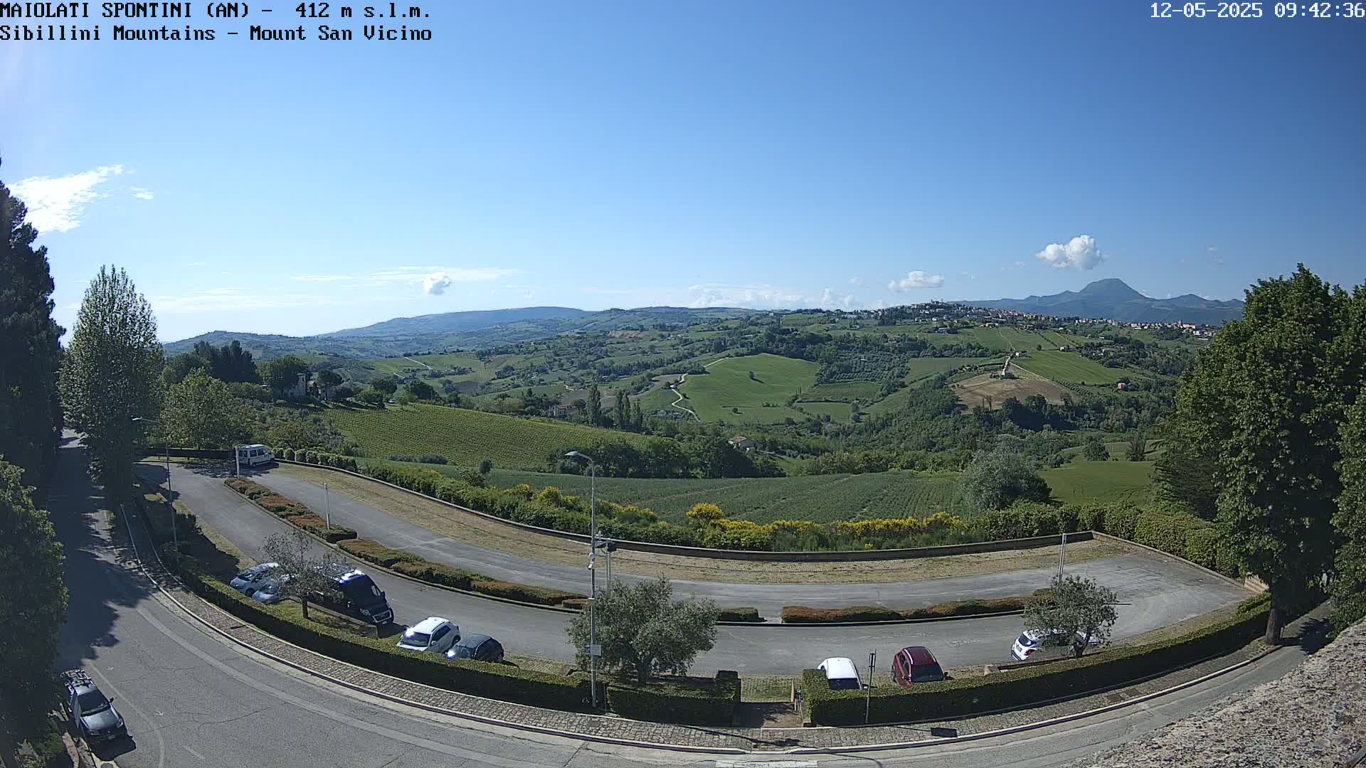 A curved road with parked cars overlooks a sunny, green valley and distant mountains under a clear blue sky.