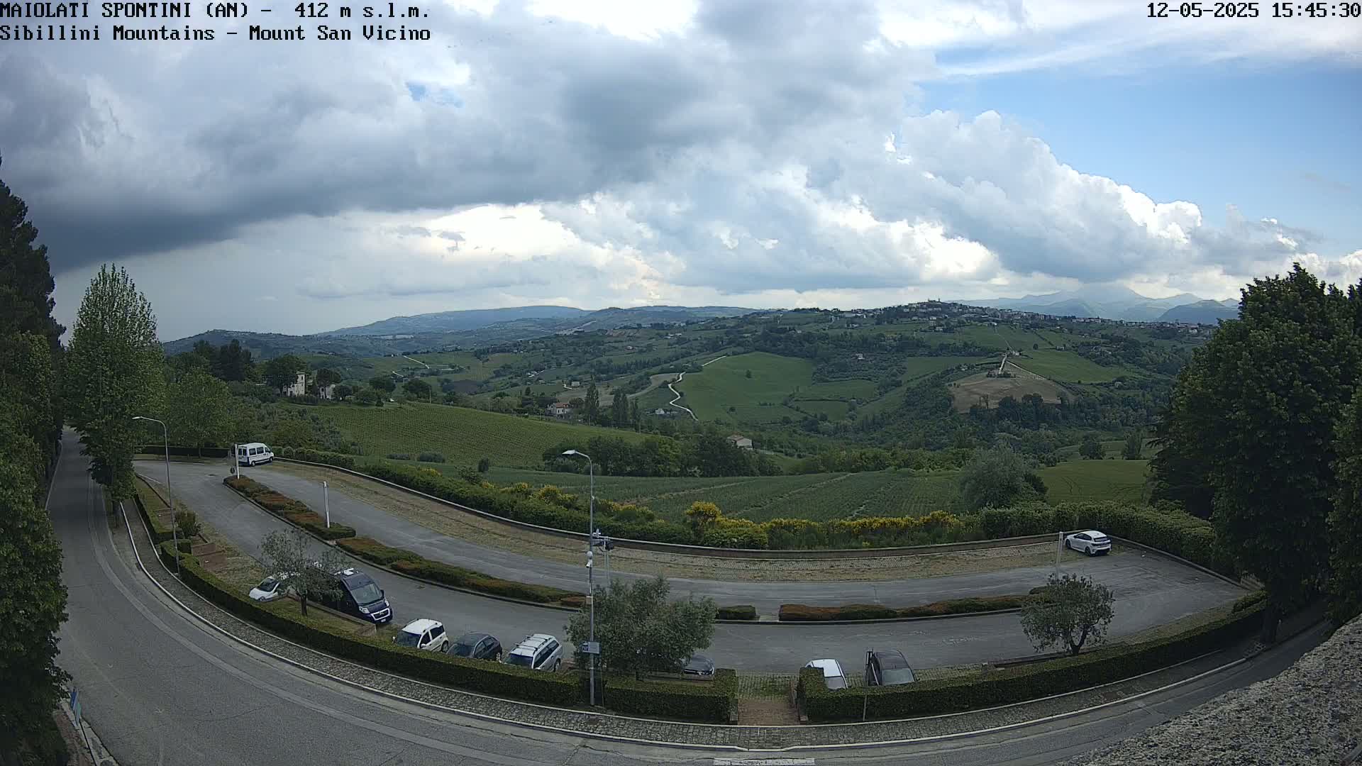 A partly cloudy sky overlooks a hilly landscape of green fields and vineyards, with a curved road and a parking lot containing several cars.