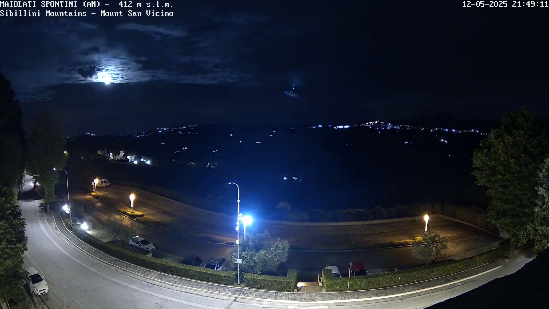 A nighttime view of a curved road with parked cars, illuminated by streetlights, overlooks a distant town nestled in hills under a partly cloudy sky and bright moon.