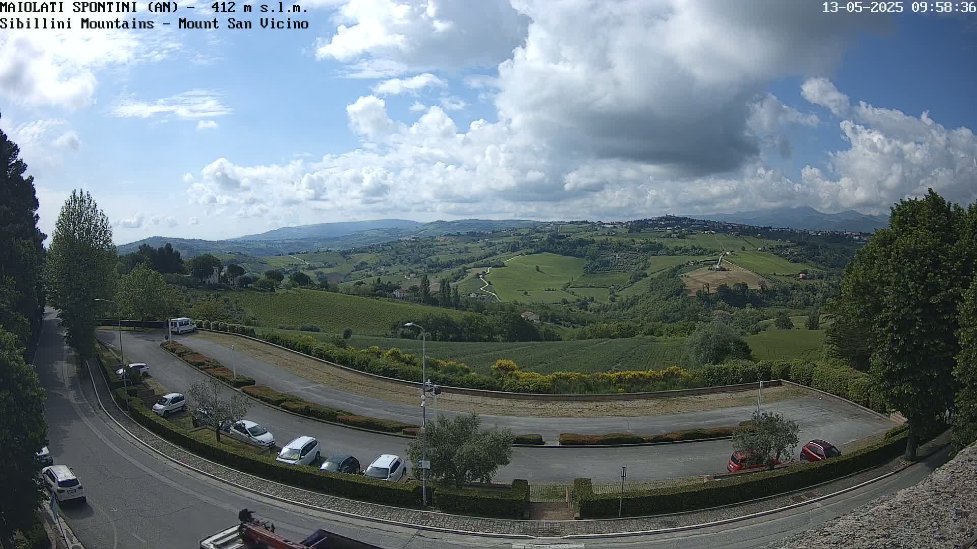 A partly cloudy sky overlooks a hilly landscape with vineyards and a parking lot containing several cars.