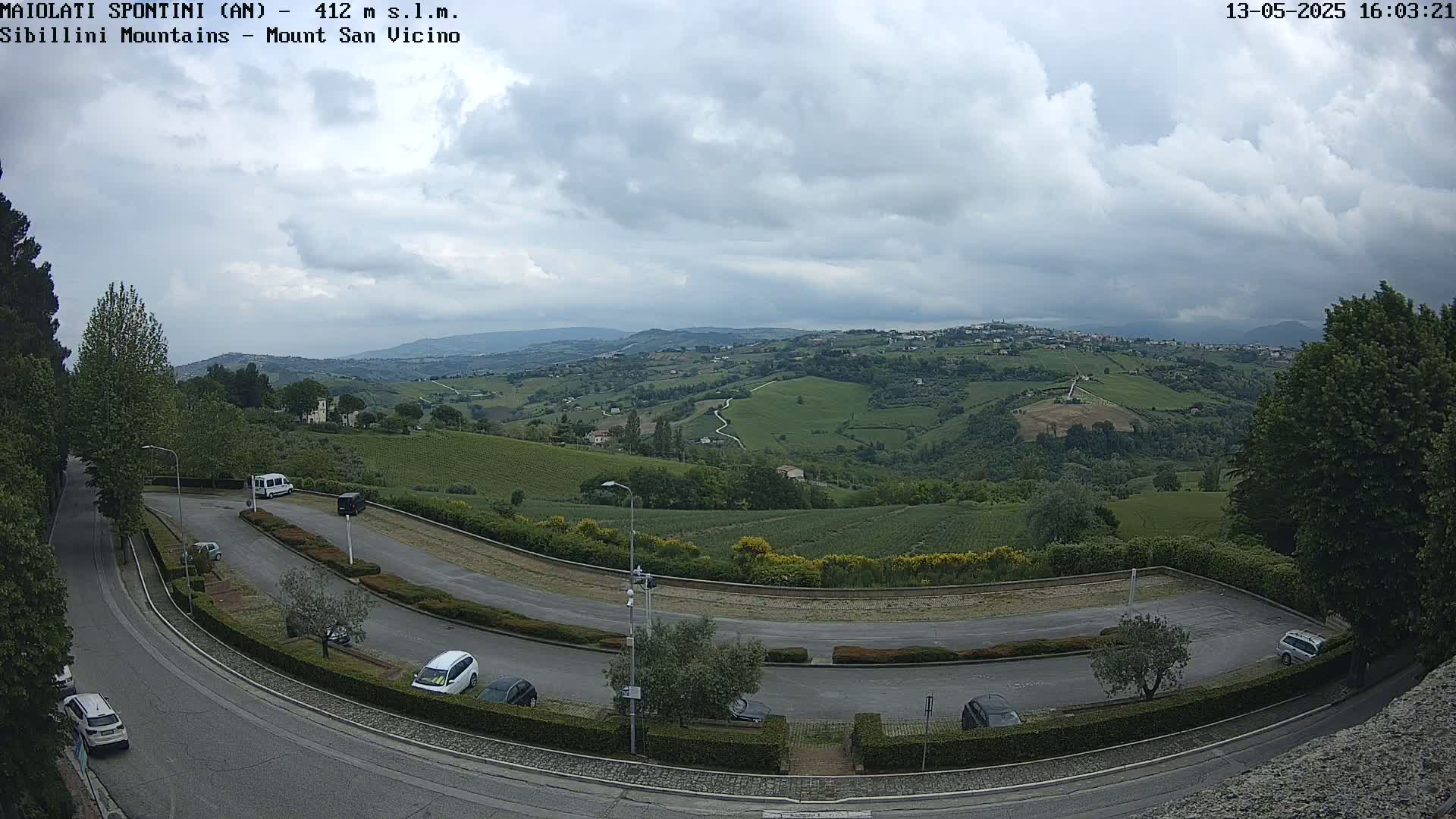 A mostly cloudy day reveals a sweeping view of a hilly, green landscape dotted with buildings and vineyards, overlooked by a curved roadway with parked cars.