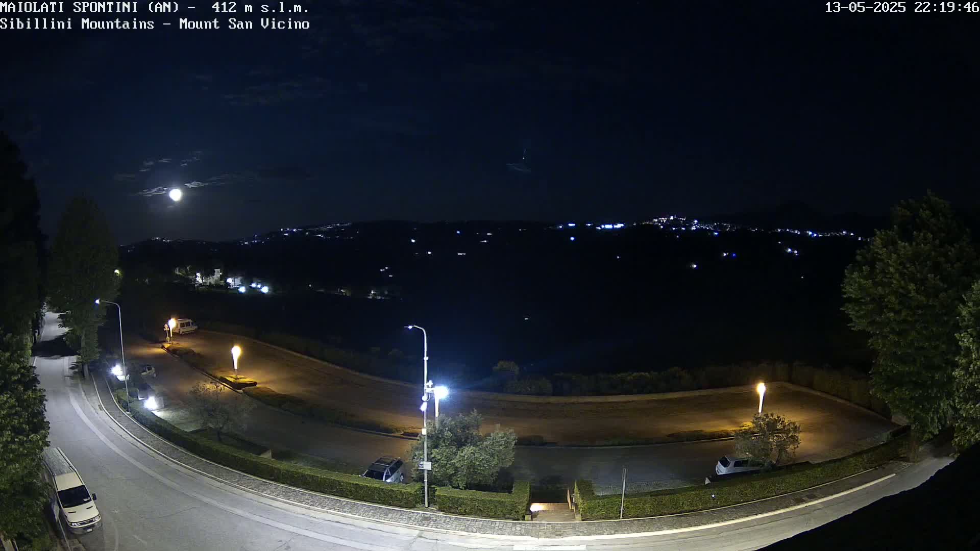 A nighttime view of a curving road with parked cars and illuminated light posts, overlooking a dark, hilly landscape under a bright moon.