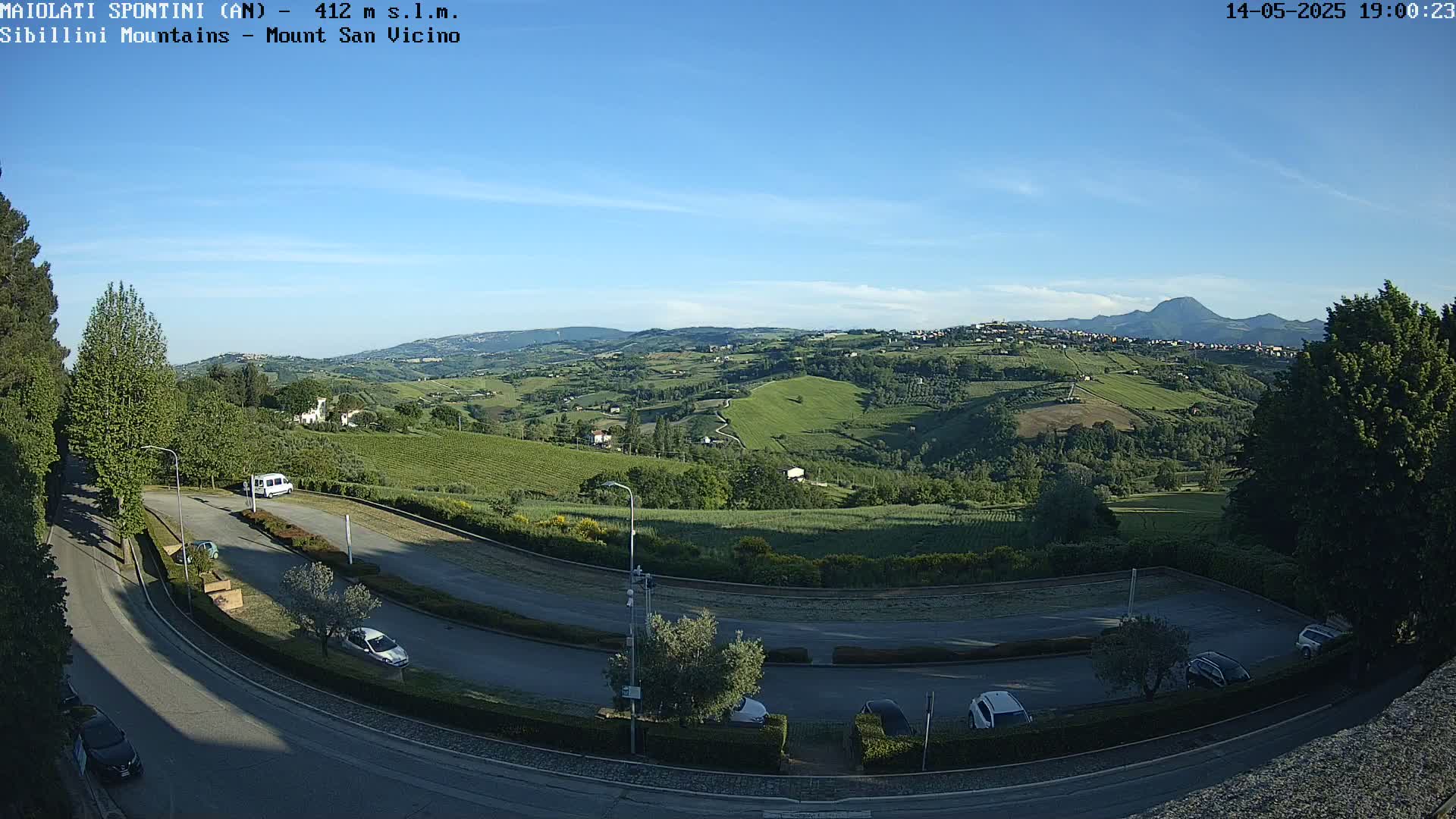 A high-angle view shows a sunny, clear day overlooking a hilly landscape with vineyards and a parking lot containing several cars; a mountain range is visible in the distance.