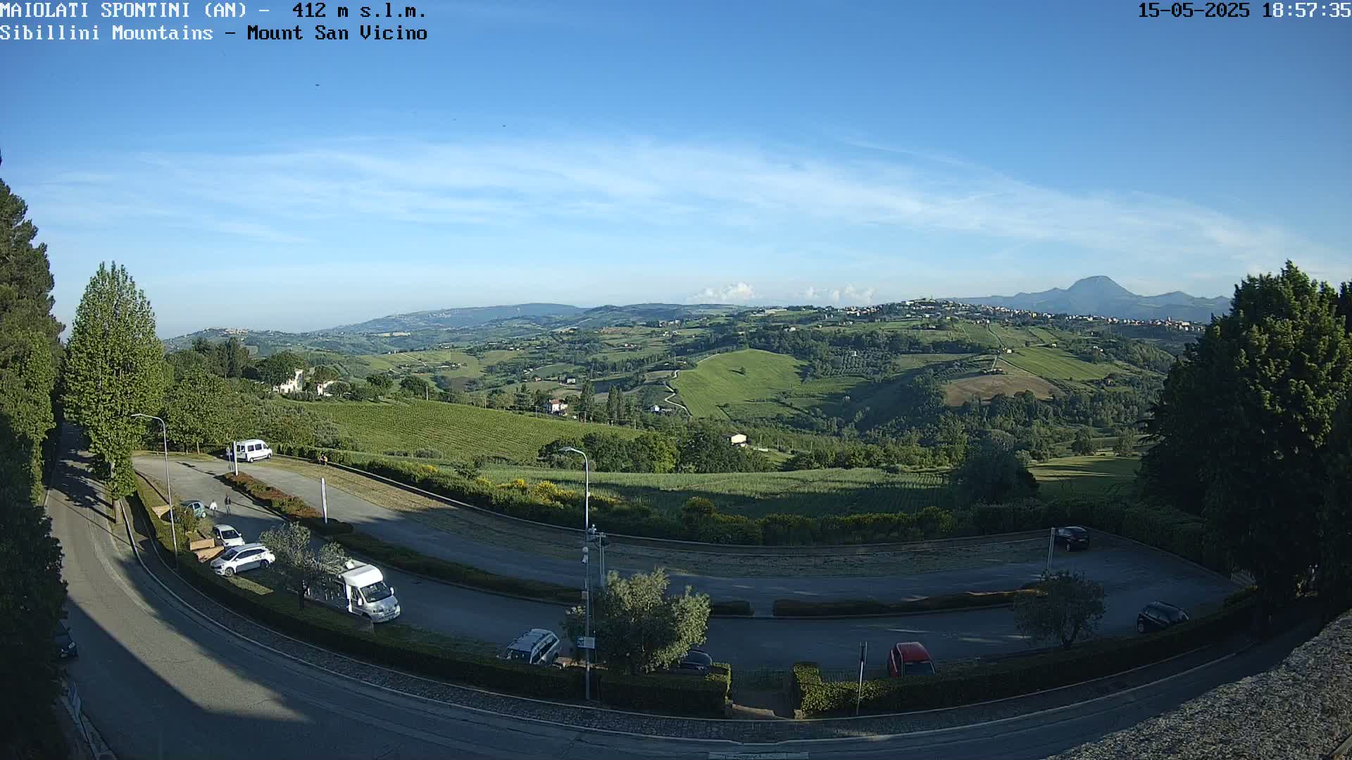 A winding road curves through a parking lot overlooking a sunlit valley of rolling green hills and distant mountains under a clear blue sky.