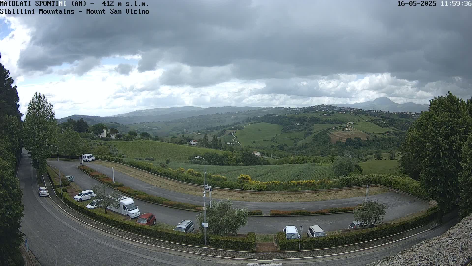 A parking lot with several cars and vans overlooks a hilly landscape of green fields and vineyards under a cloudy sky.