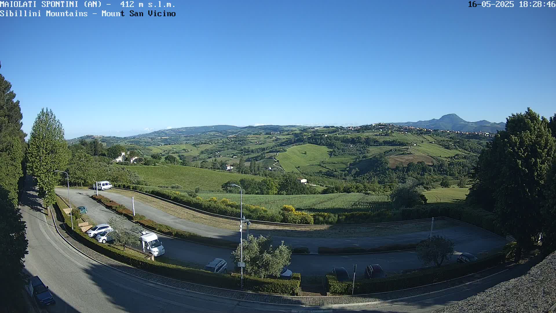 A high-angle, long shot shows a sunny, clear day overlooking a hilly, green landscape with a parking lot and road in the foreground.
