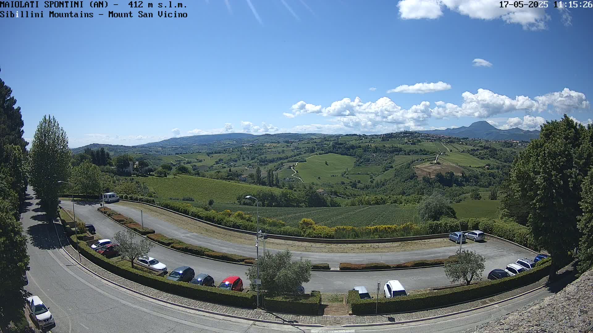 A partly cloudy day reveals a hilly landscape with vineyards and a parking lot, observed from an elevated vantage point.